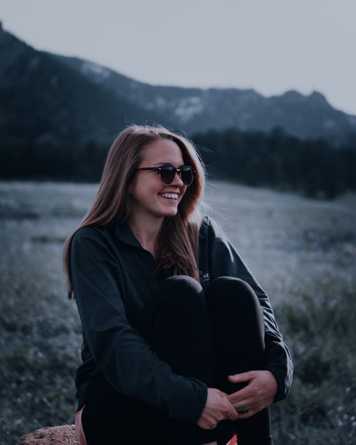 Happy woman with sunglasses sitting outdoors in a field with a mountain backdrop, embracing nature's serenity.