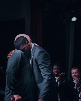 舞台上，两个穿着西装的男人情绪激动地拥抱在一起，周围是鼓掌的观众