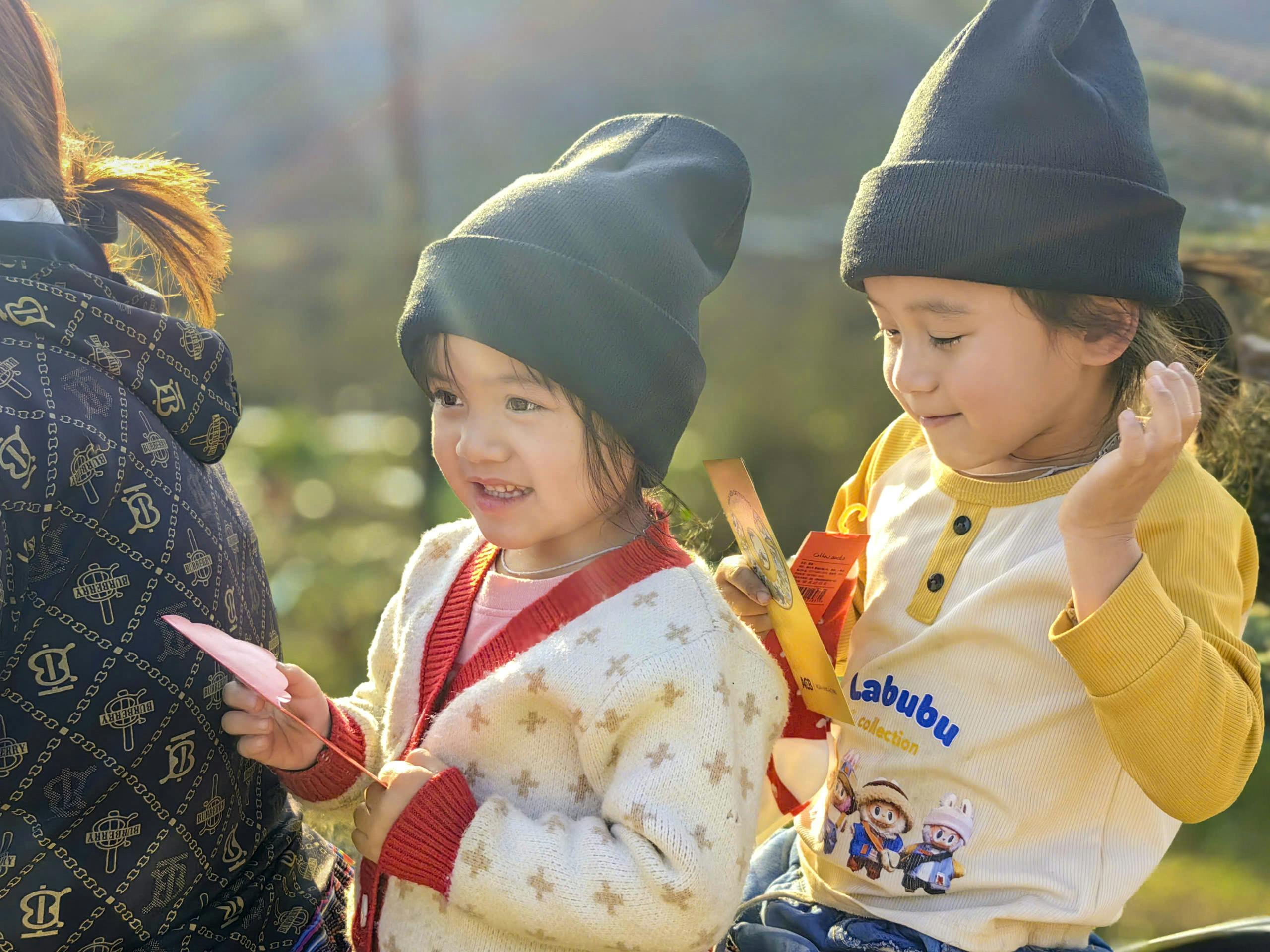 Two children in beanies enjoying a sunny day, holding cards and playing.