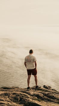 A solitary figure stands on a rocky cliff gazing out at a calm sea under a pale sky.