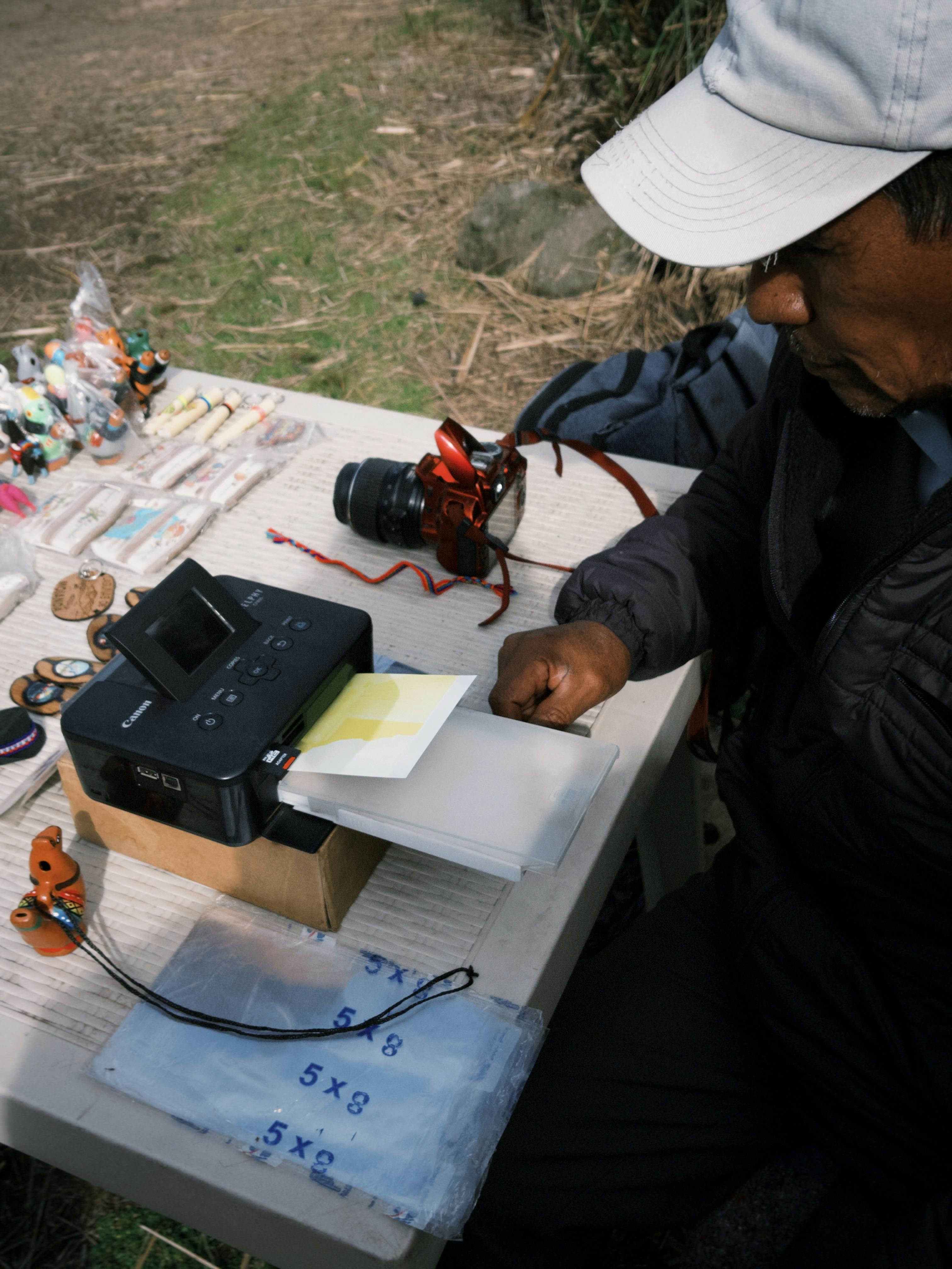 Vendor printing photos with portable printer at outdoor market stall, showcasing handmade crafts.