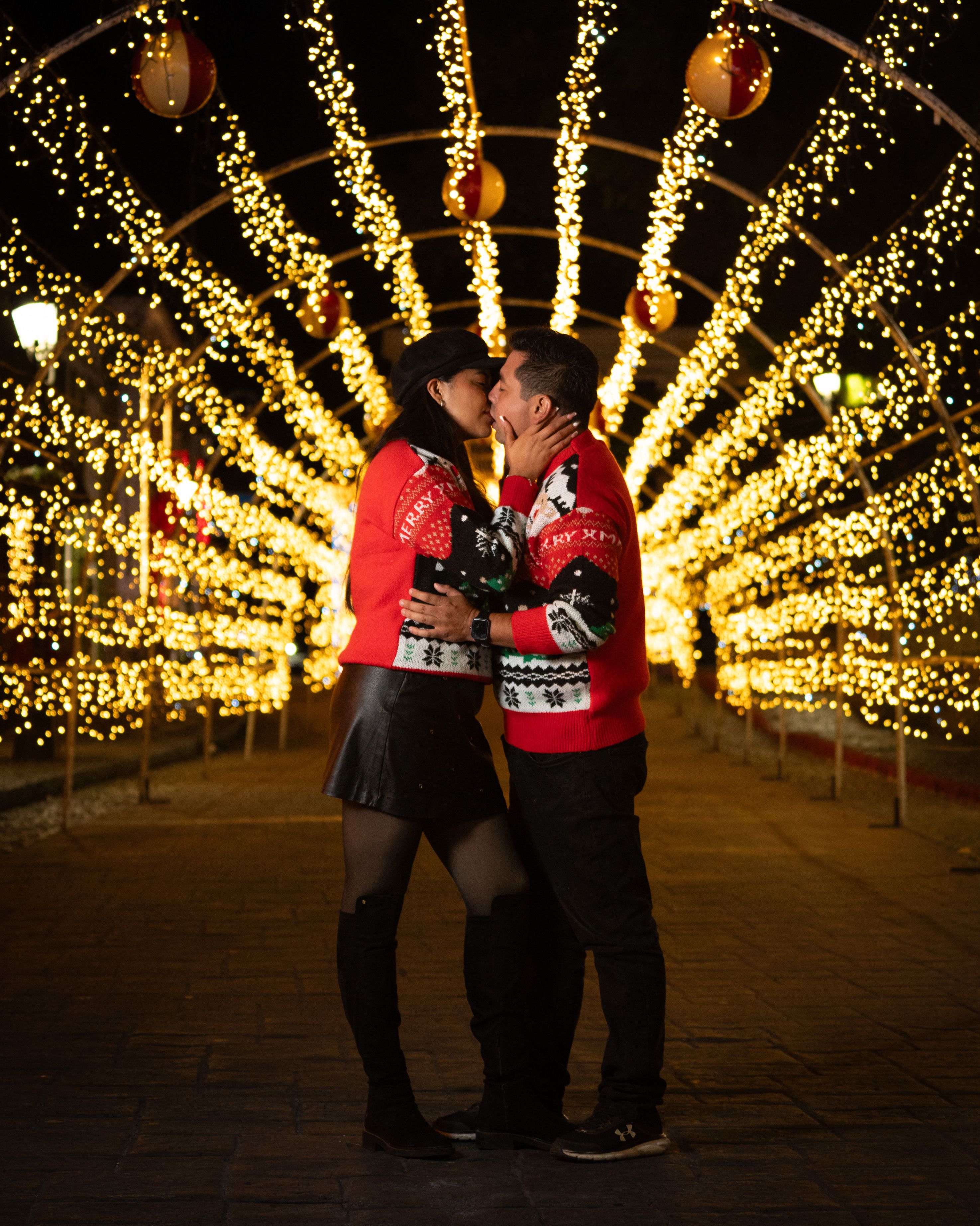 Couple in red sweaters kissing under illuminated Christmas decorations at night.