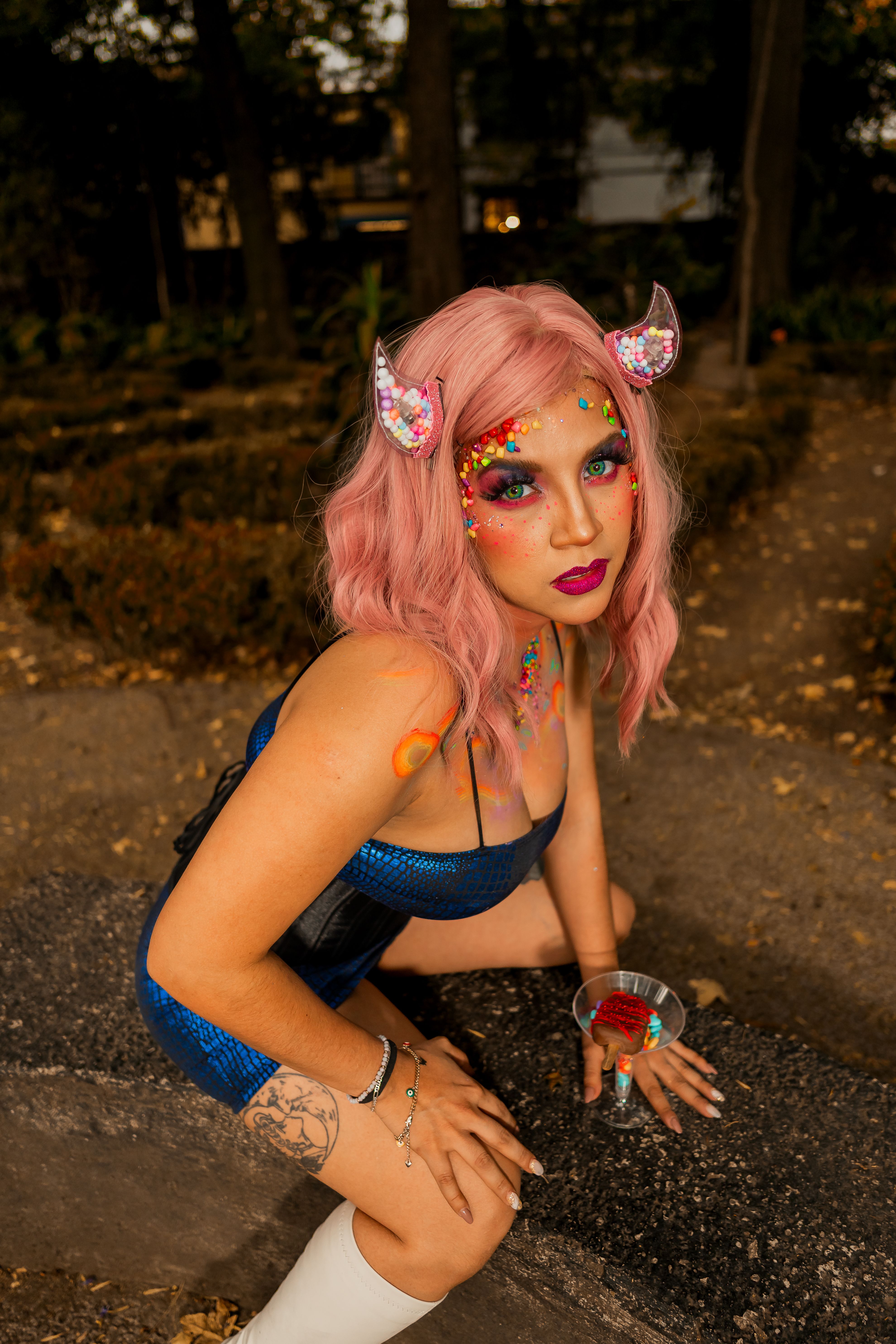 A woman with surreal makeup and costume poses outdoors, blending fantasy and vibrant colors.