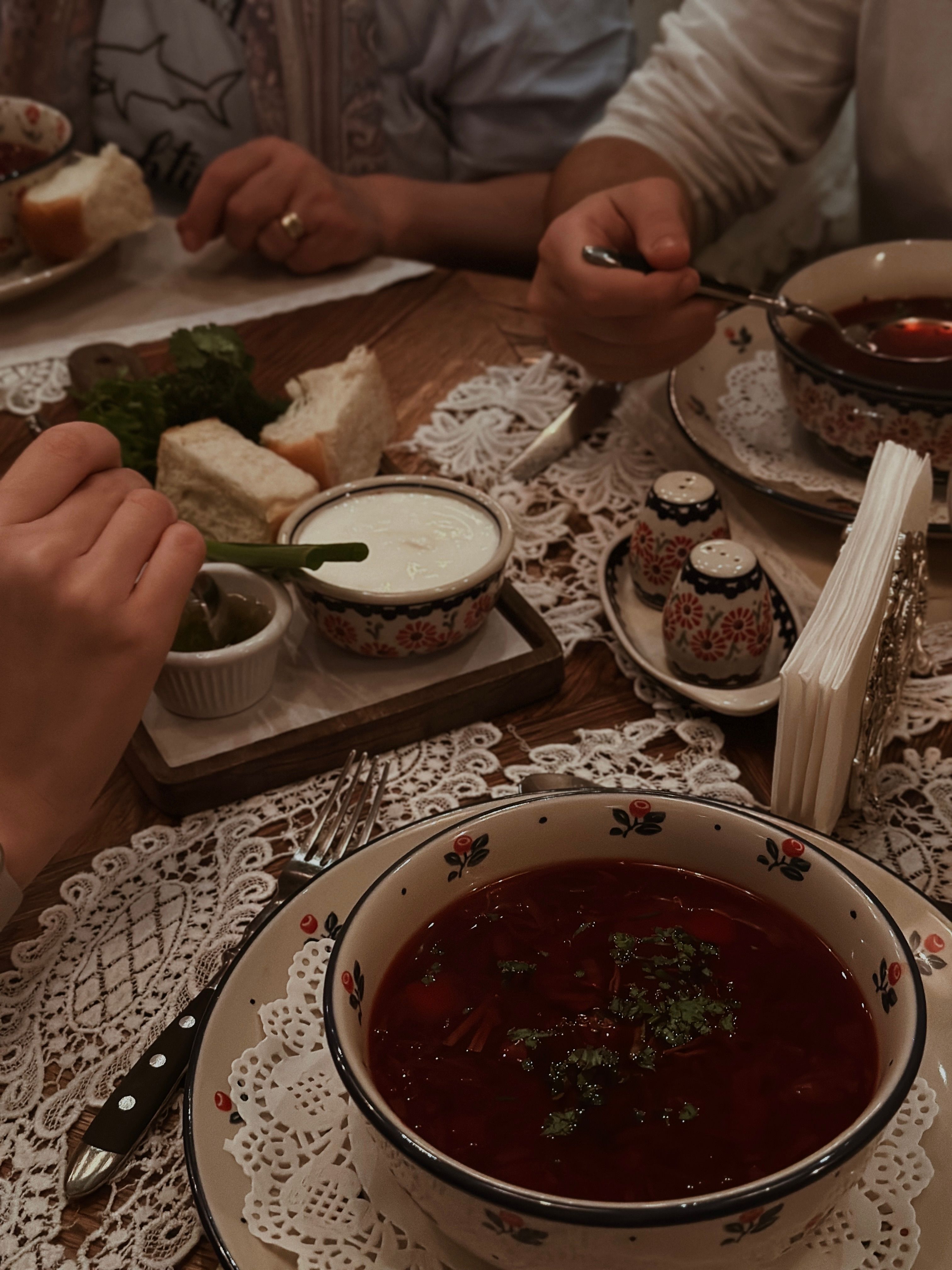 Table nappée de dentelle : bol de bortsch au premier plan, pot de crème, pain tranché et salières à motifs floraux rouges et bleus, une main au bord du cadre