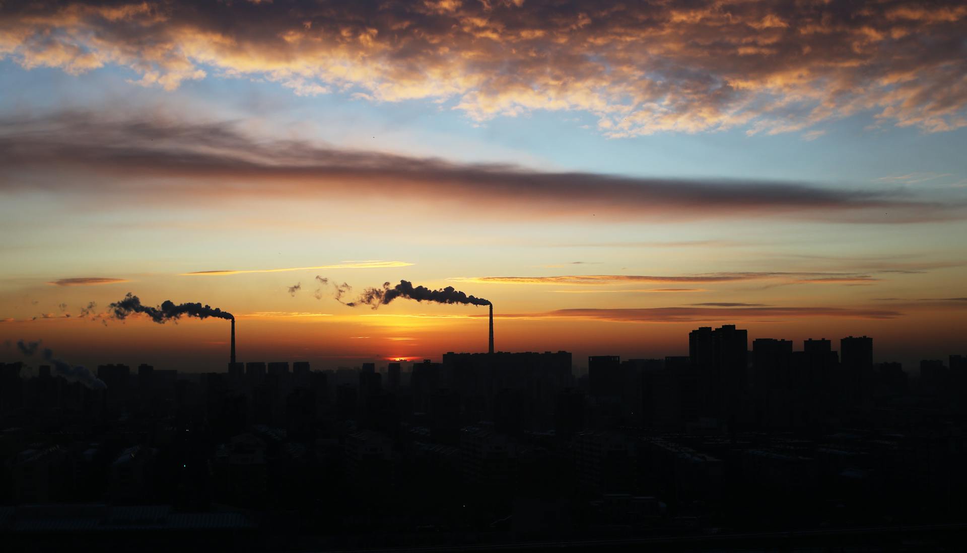 A dramatic city skyline at sunset with smokestacks emitting pollution into the sky.