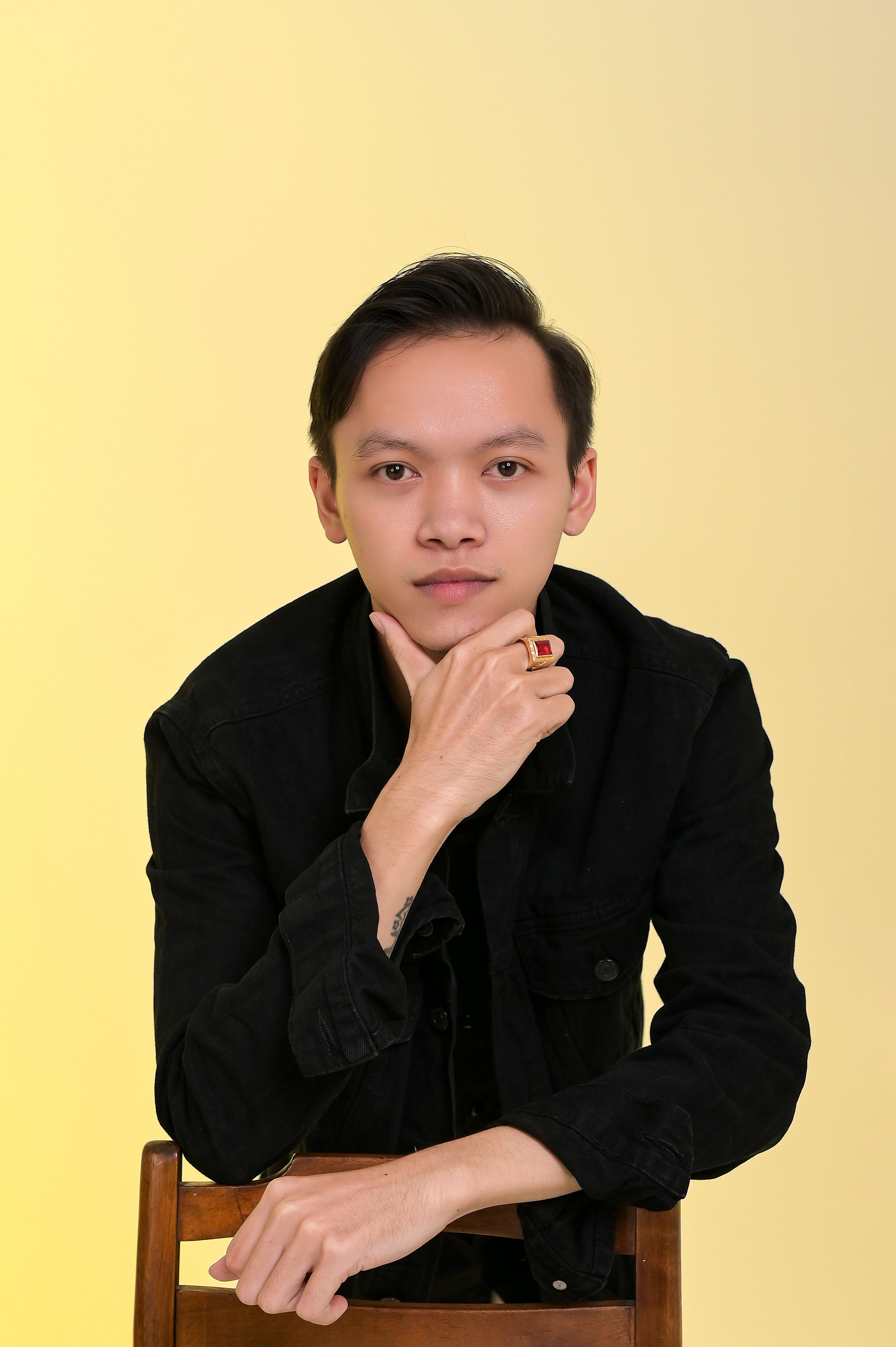 A young man in a black jacket poses thoughtfully against a yellow background.