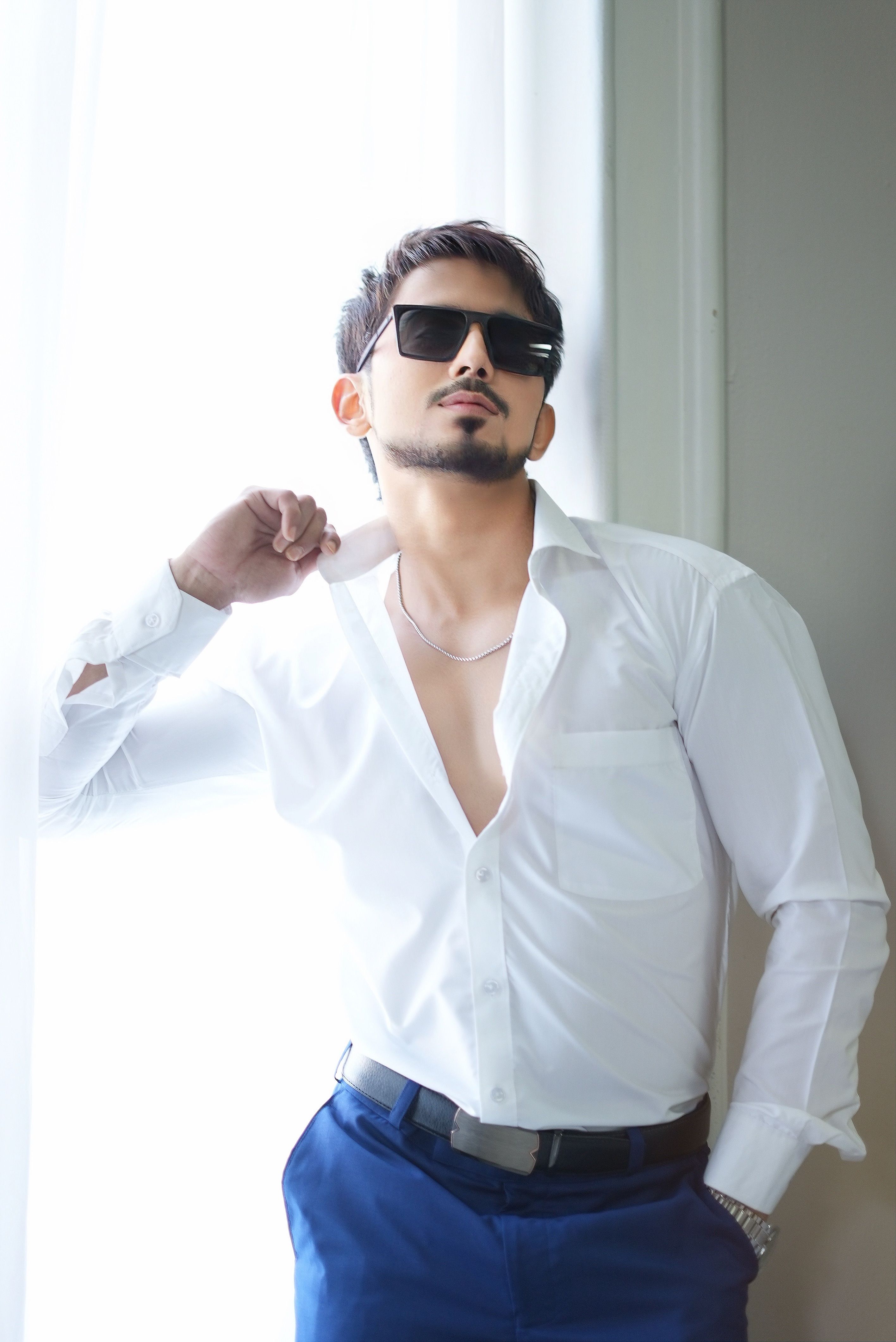 Confident man in sunglasses posing by a bright window, exuding modern style.