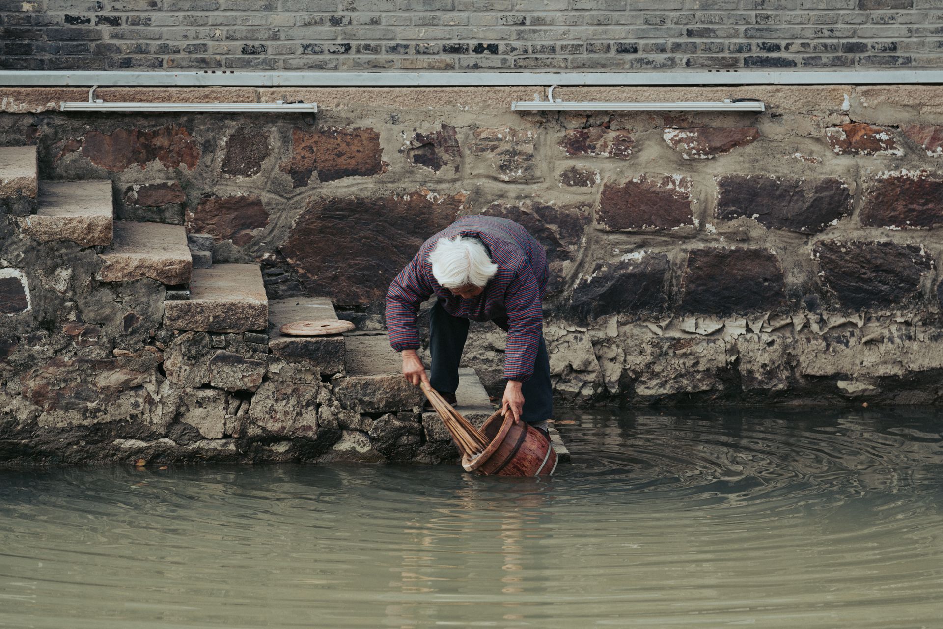 An elderly person gathering water from a pond beside a rustic stone wall and stairs.