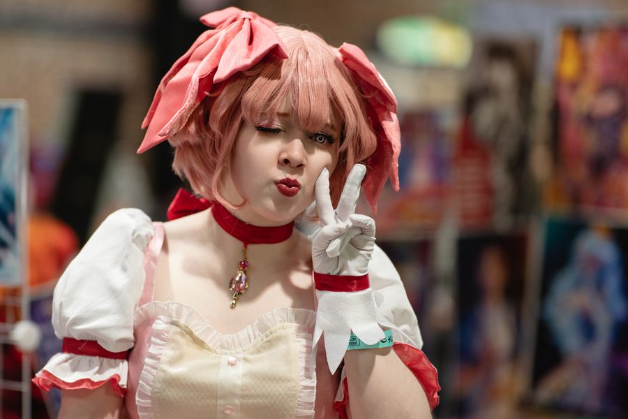 Cosplayer in pink wig and dress posing with peace sign at an indoor convention hall with posters in background