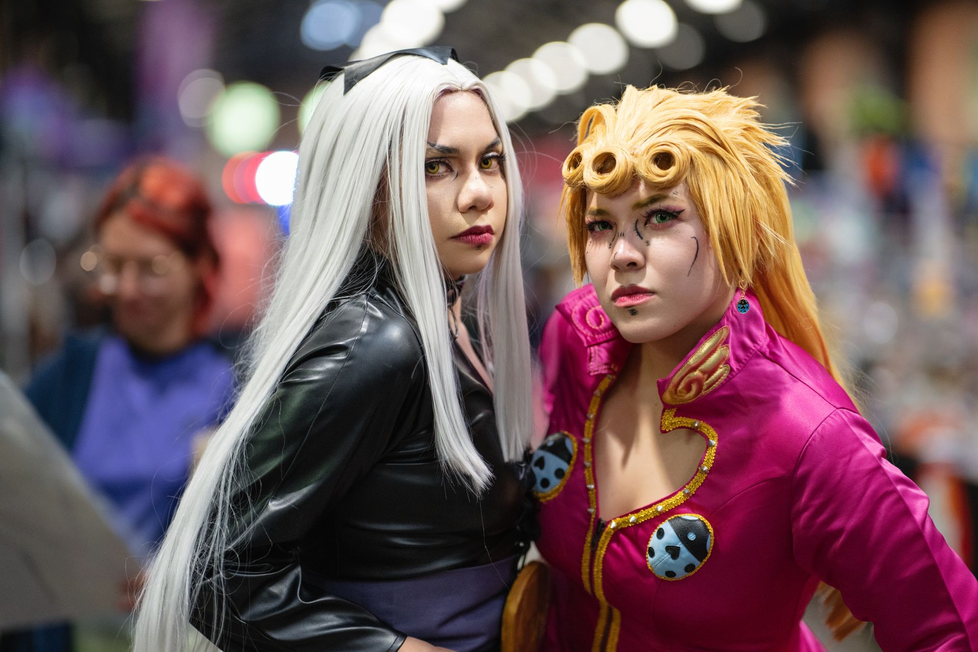 Two cosplayers in elaborate costumes and wigs pose at a comic convention show floor