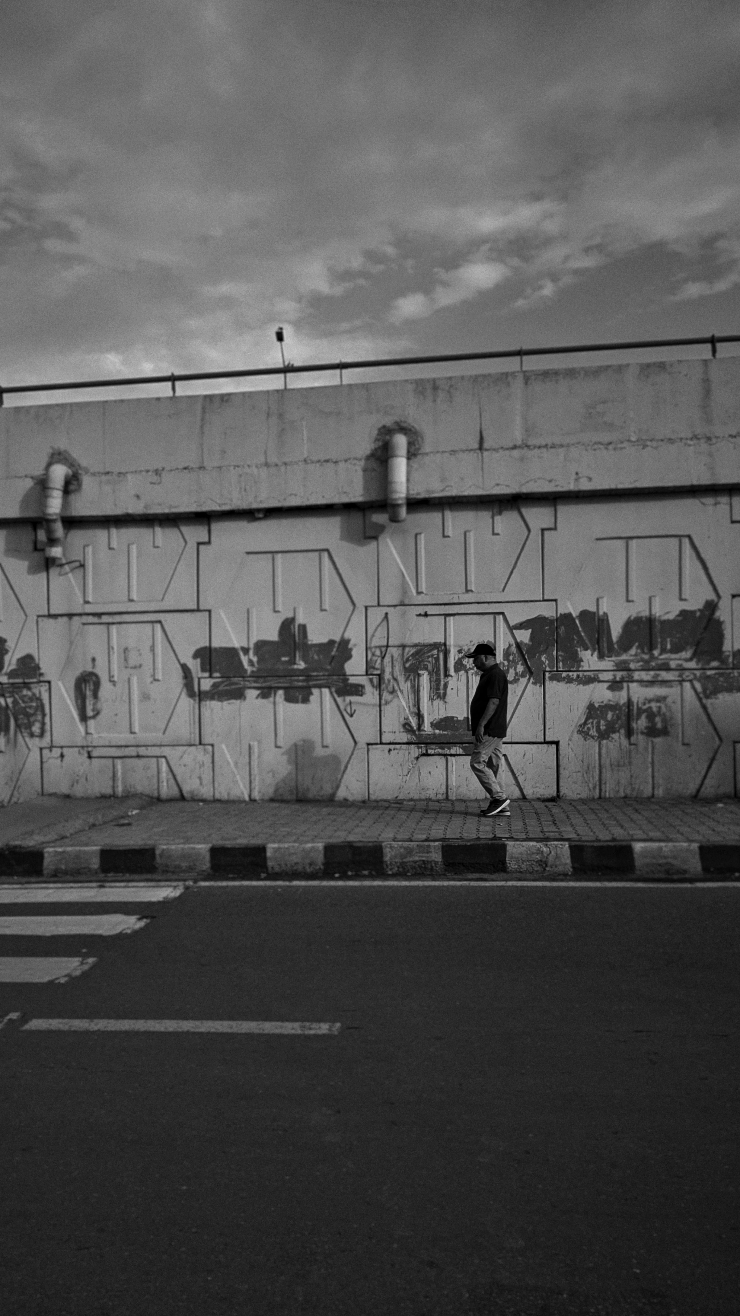 Black and white photo of a lone person walking by graffiti on a street wall.