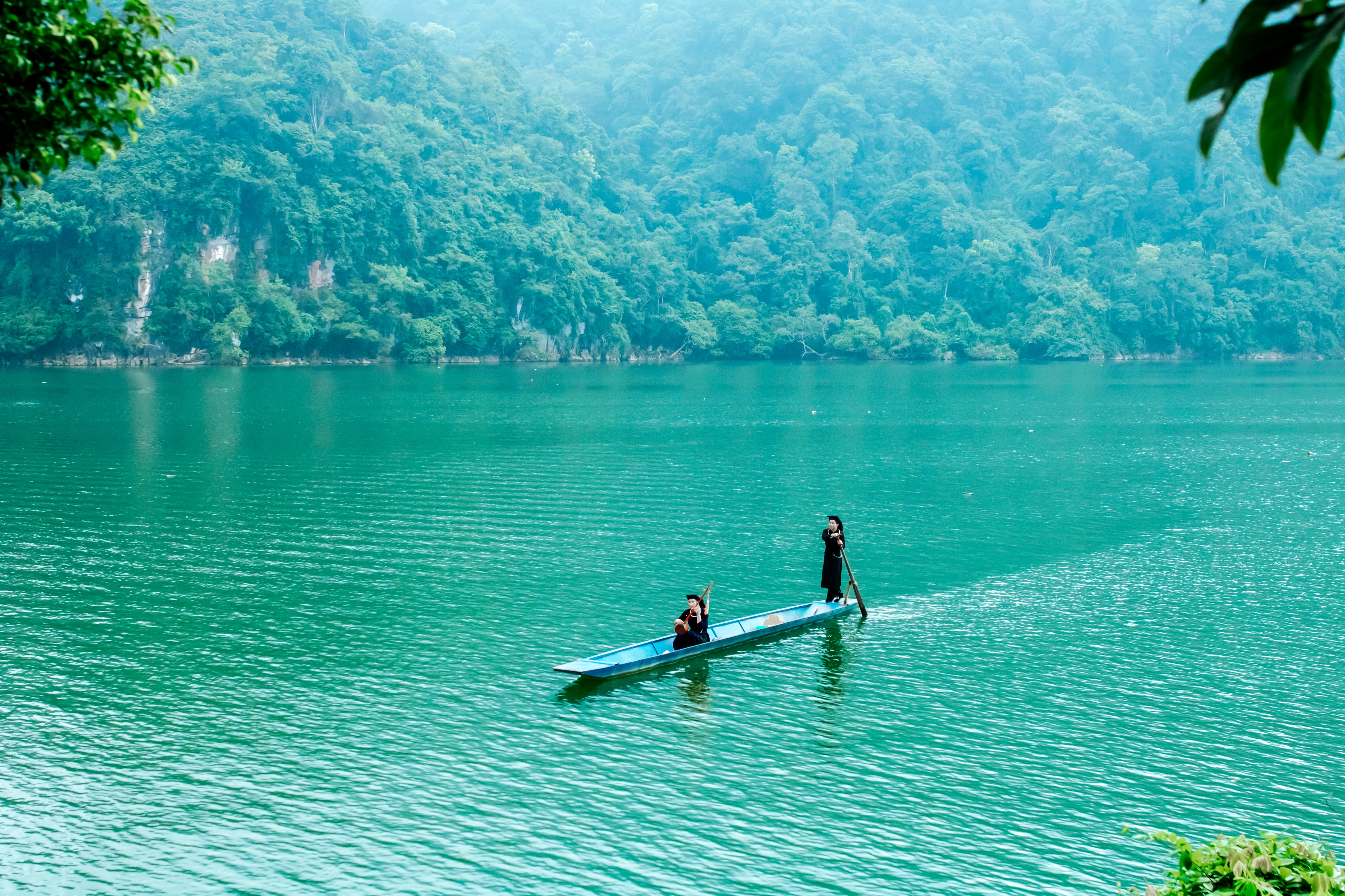 Two people in a boat navigate a tranquil, emerald-colored lake surrounded by lush forest.