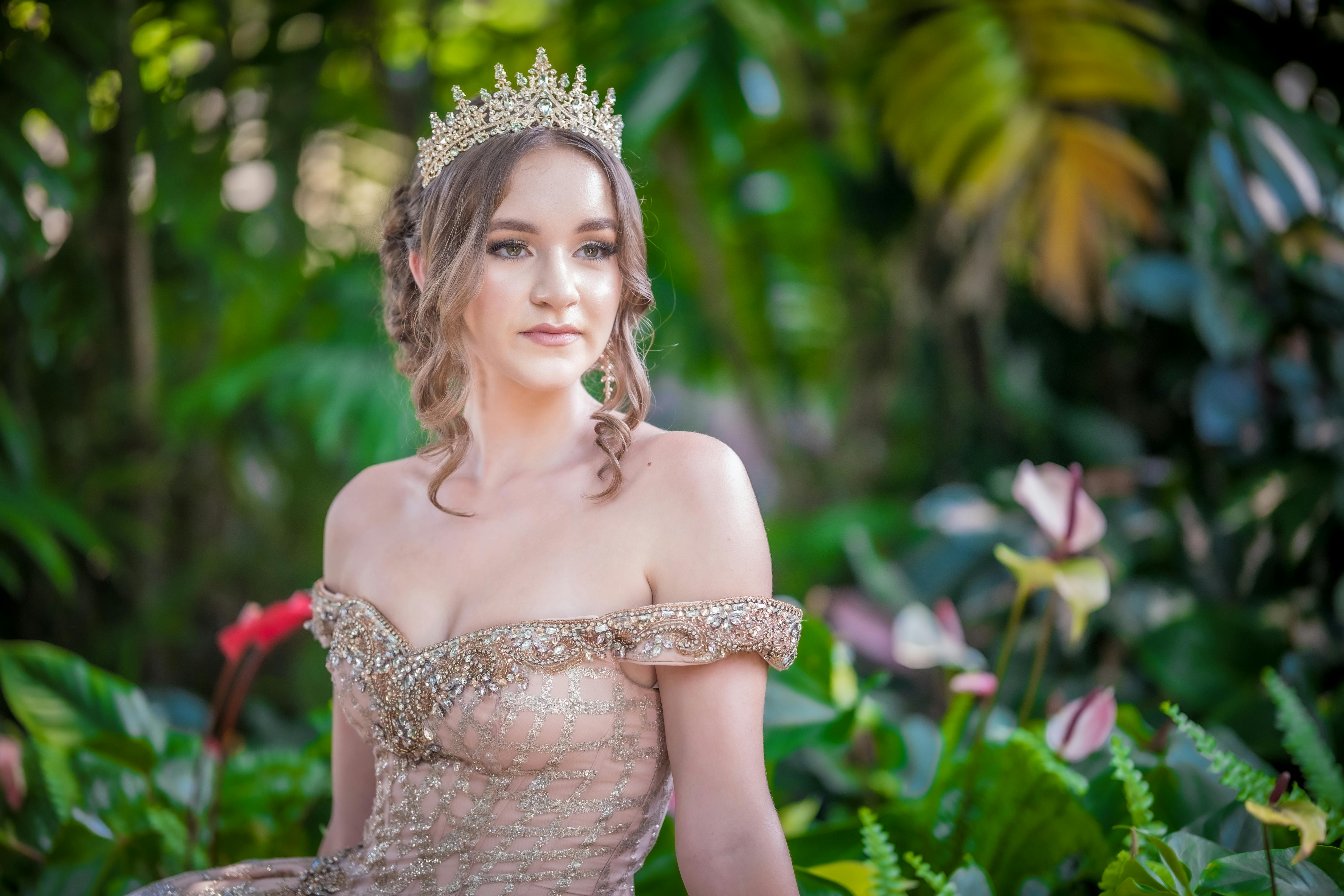 Captivating portrait of a woman in a shimmering gown and tiara amidst lush greenery.