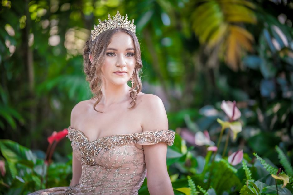 Captivating portrait of a woman in a shimmering gown and tiara amidst lush greenery.