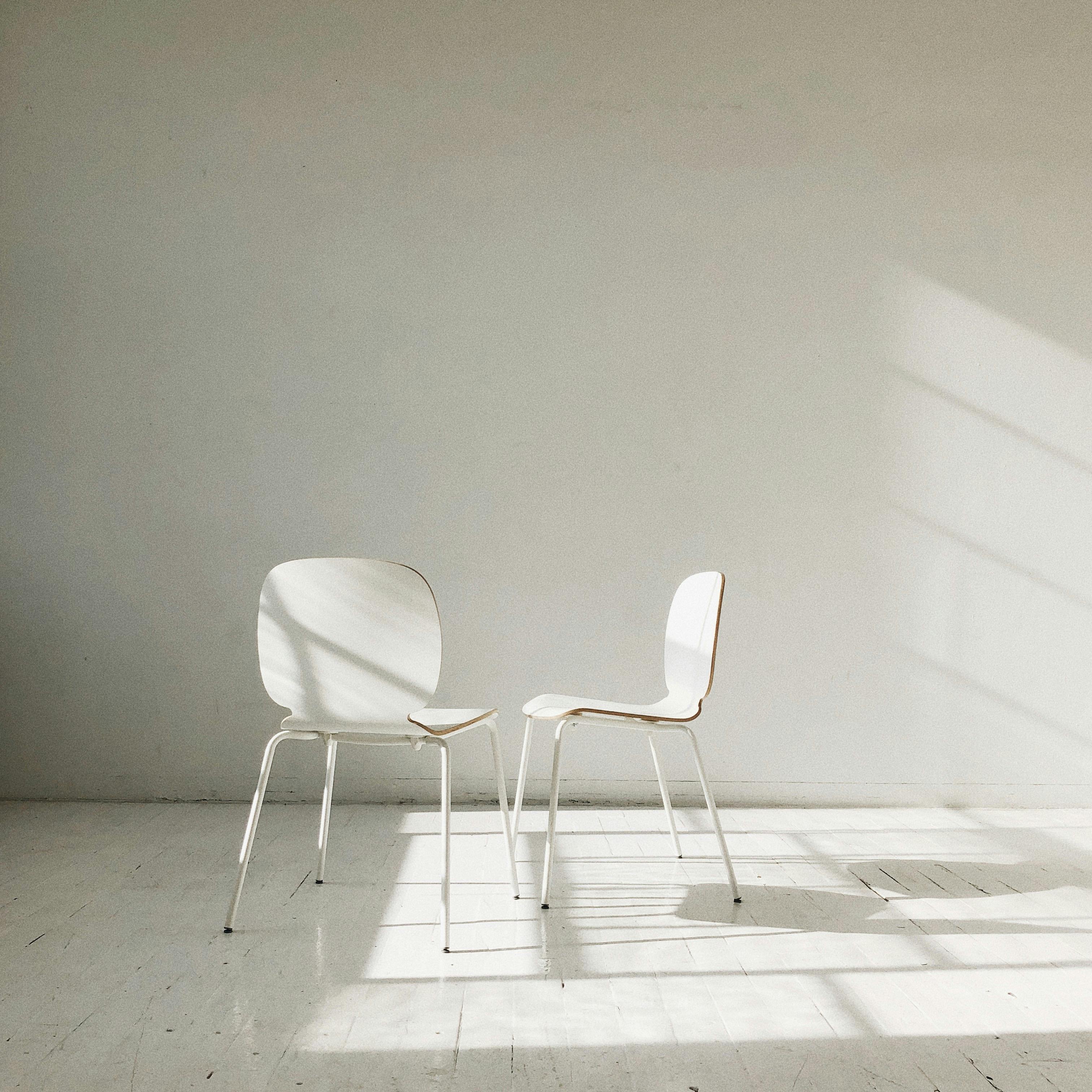 Two modern white chairs in a sunlit, minimalist interior with soft shadows.