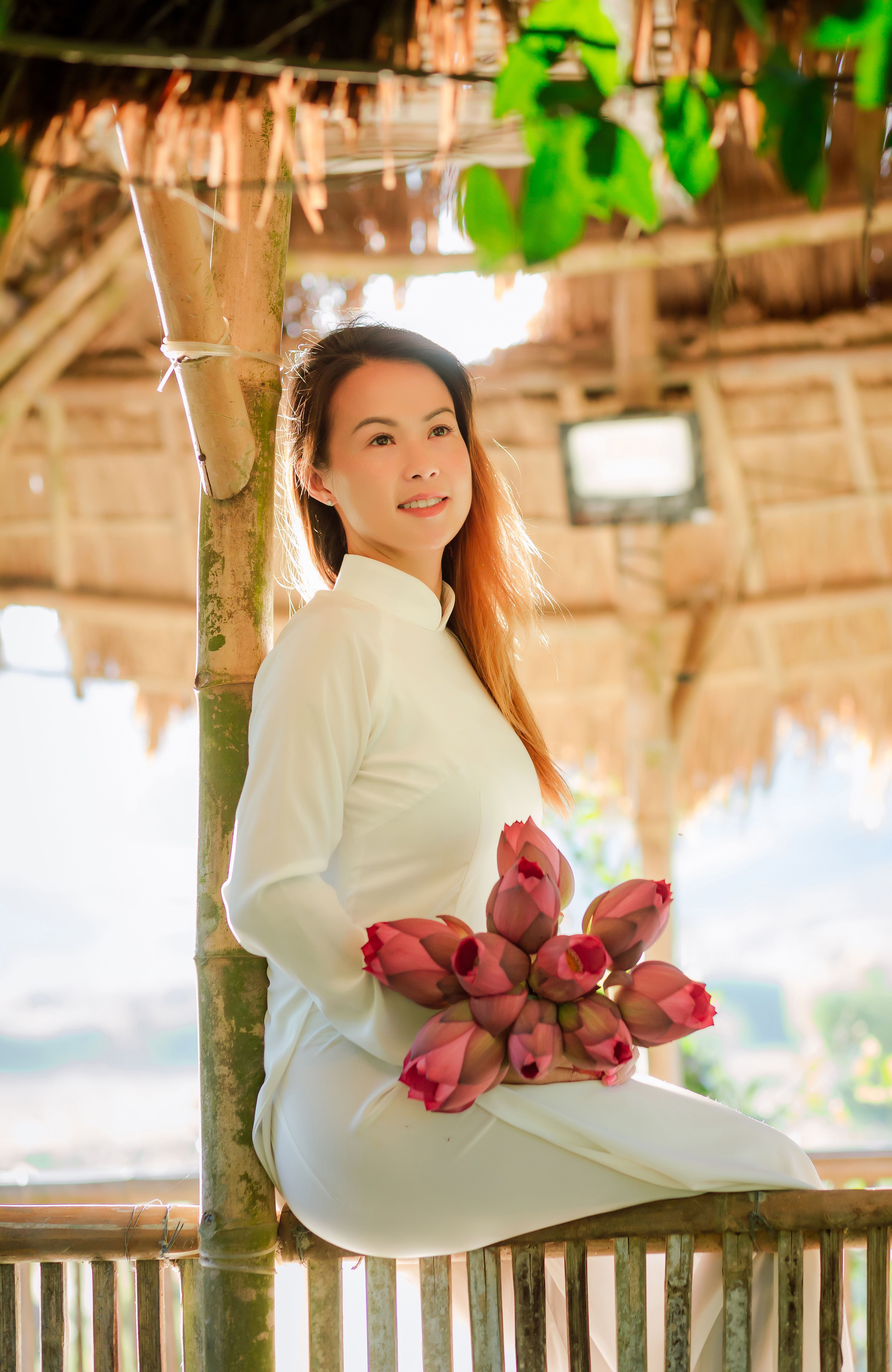 A woman in a white Ao Dai holding lotus flowers in a serene Vietnamese setting.