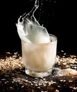 A dramatic shot of oat milk splash in a glass against a black background with scattered oats.