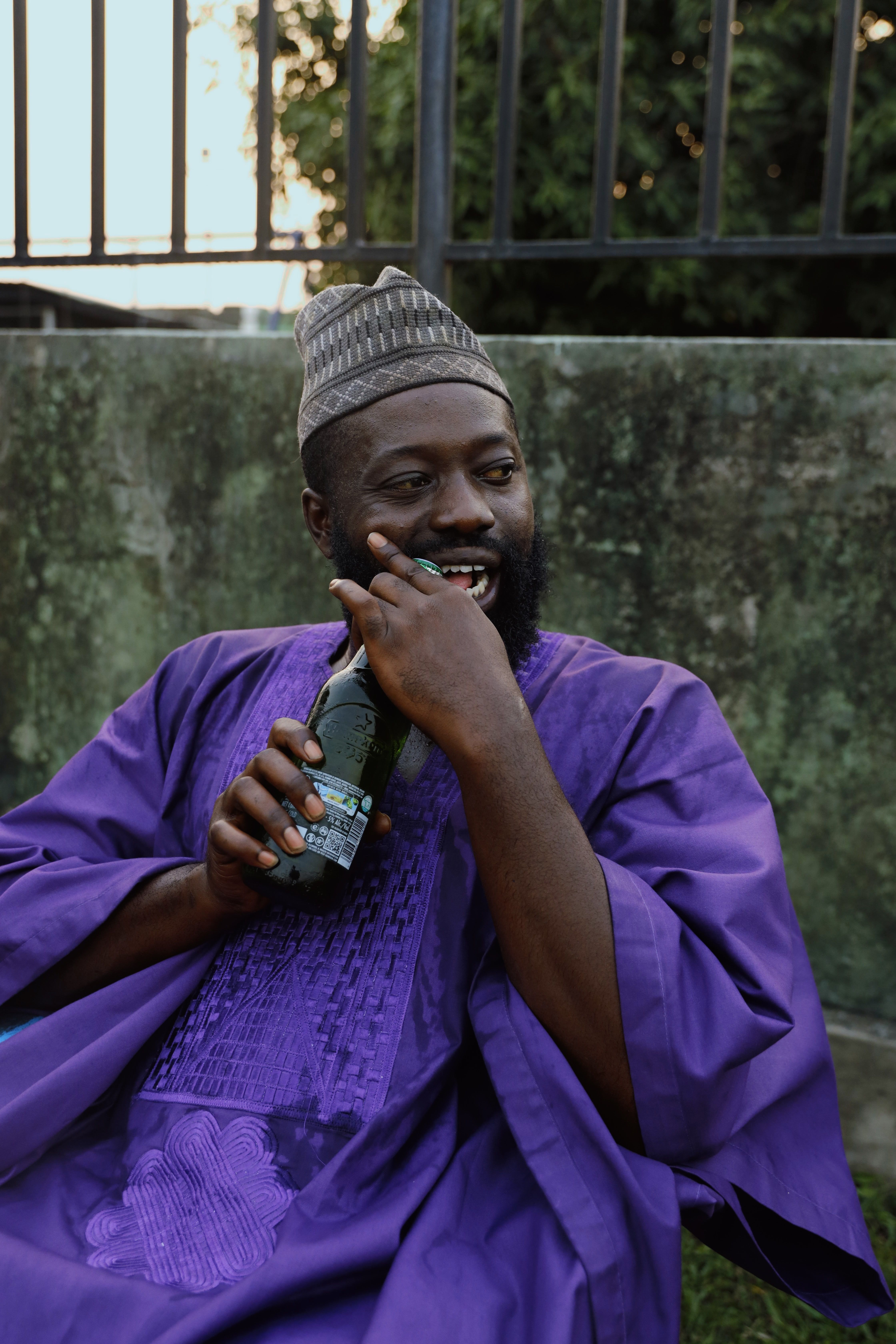 Man in a purple traditional outfit seated outdoors, holding a drink and smiling.