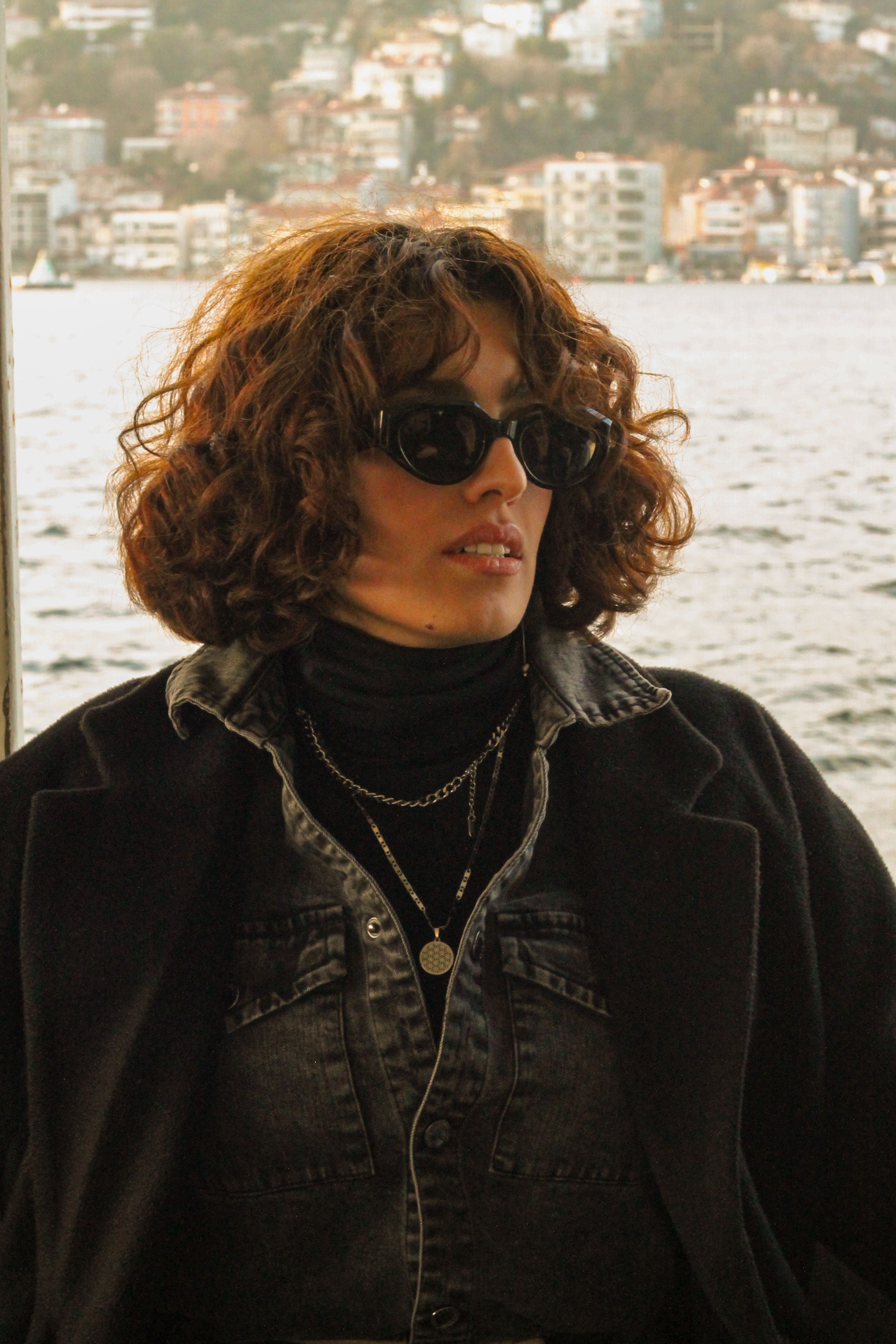 Woman in sunglasses and black coat with curly hair poses by a city waterfront, exuding modern elegance.