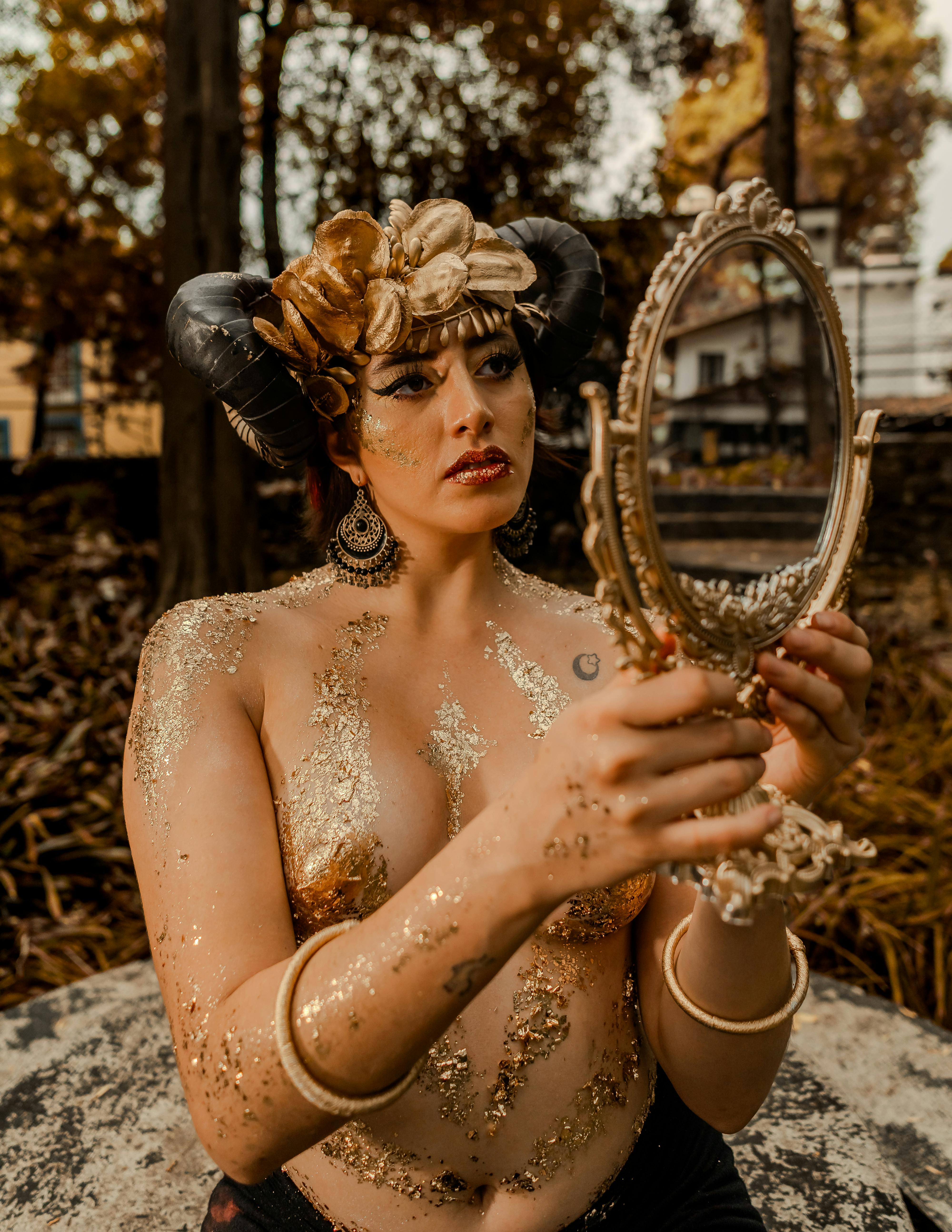 grátis Uma mulher com maquiagem surreal e chifres segura um espelho decorativo em um ambiente externo, criando uma cena artística e fantástica. Foto profissional