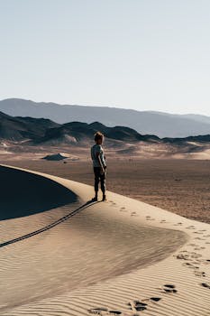 蔚蓝的天空下，一位孤独的女人站在沙丘顶上，背景是山脉
