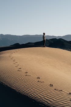阳光明媚的沙丘上，一个孤独的人影站在晴朗的天空下，背景是群山