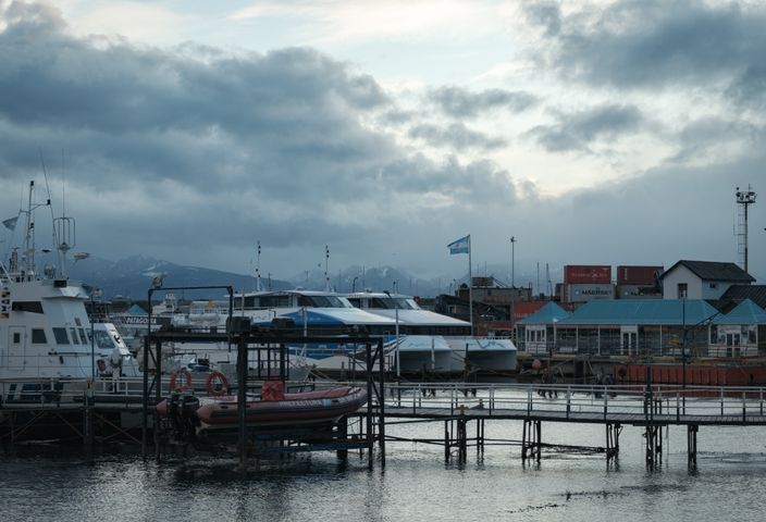 Scenic view of docked boats at Ushuaia harbor with mountain backdrop and cloudy sky.