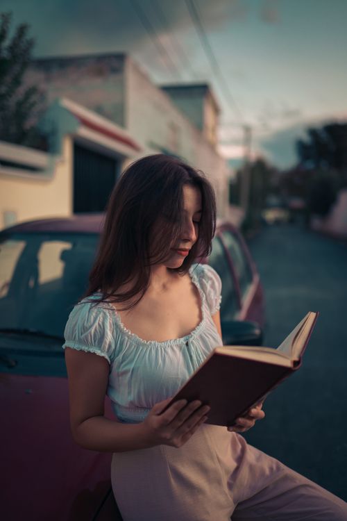 Free A young woman in casual attire reads a book outdoors against an urban backdrop at twilight. Stock Photo