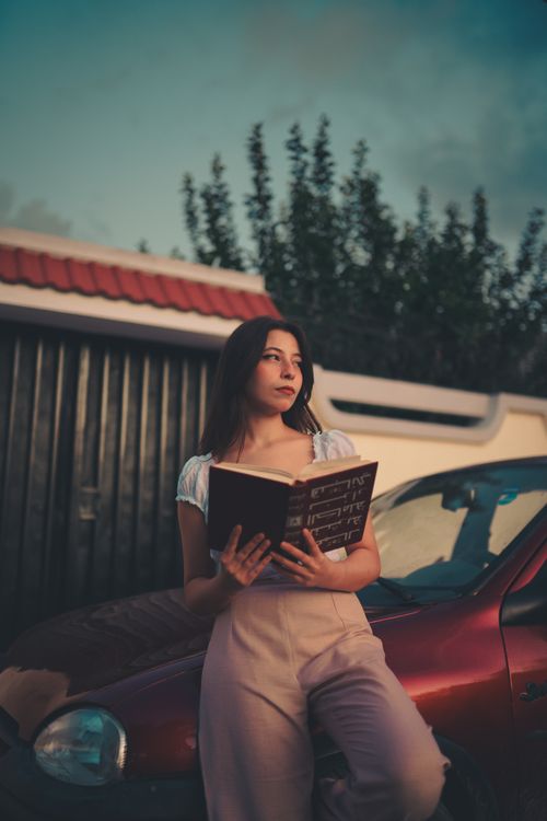 Free Young woman reading a book by a car during twilight outdoors. Stock Photo