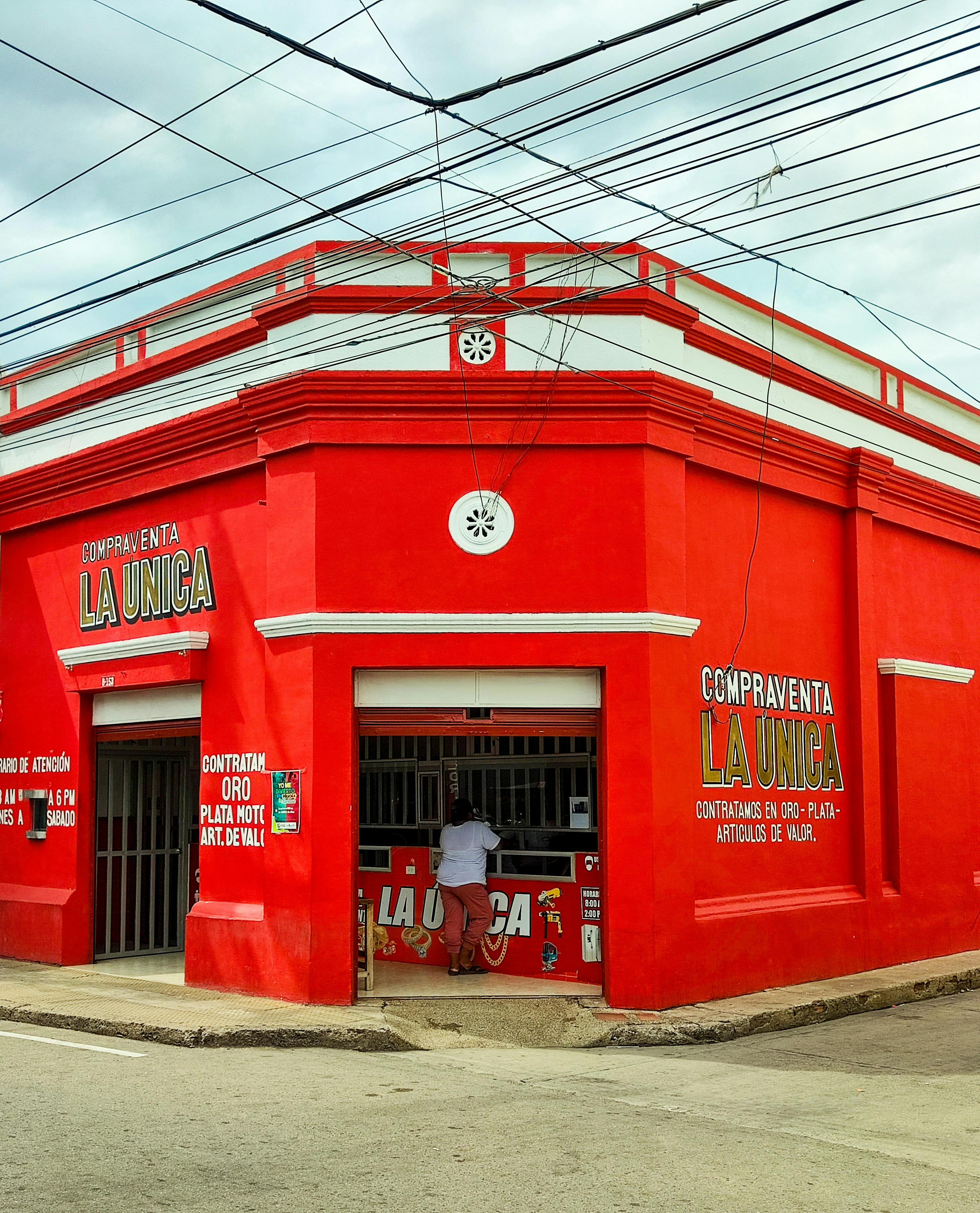 Free Street view of a bright red Compraventa La Unica storefront, featuring a person interacting inside on a cloudy day. Stock Photo