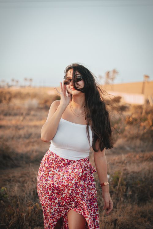 Free A woman with long hair and sunglasses stands in a sunny outdoor field, wearing a floral skirt. Stock Photo