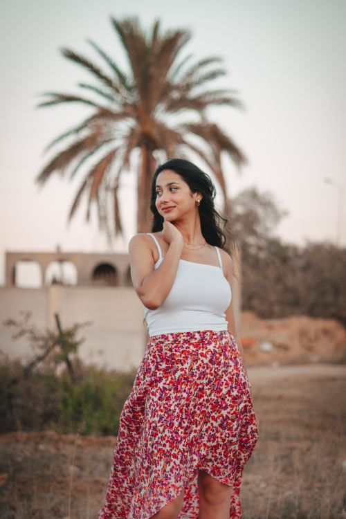 Free A young woman stands in a floral skirt with palm trees in the background, enjoying an outdoor setting. Stock Photo