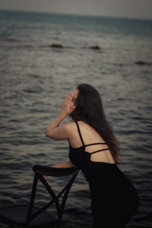 Free A woman in a black dress leans on a chair by the ocean, lost in thought at twilight. Stock Photo