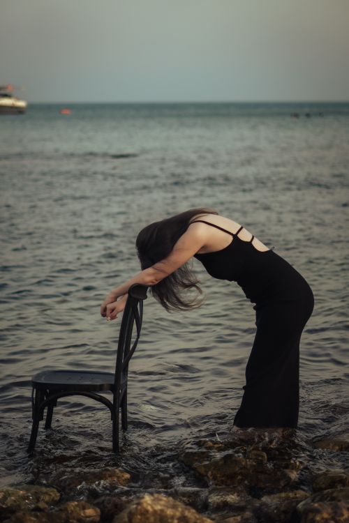 Free Artistic photo of a woman in a black dress bending over a chair on a rocky shore. Stock Photo