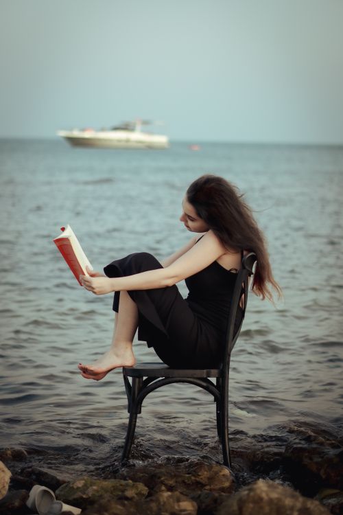 Free A woman in a black dress sits on a chair reading by the sea as a boat sails in the background. Stock Photo