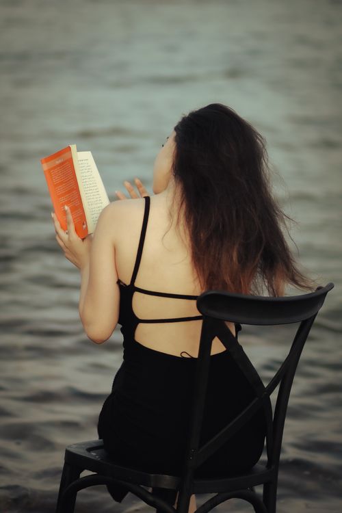 Free A woman in a black dress sits on a chair by the water, reading a book at dusk. Stock Photo