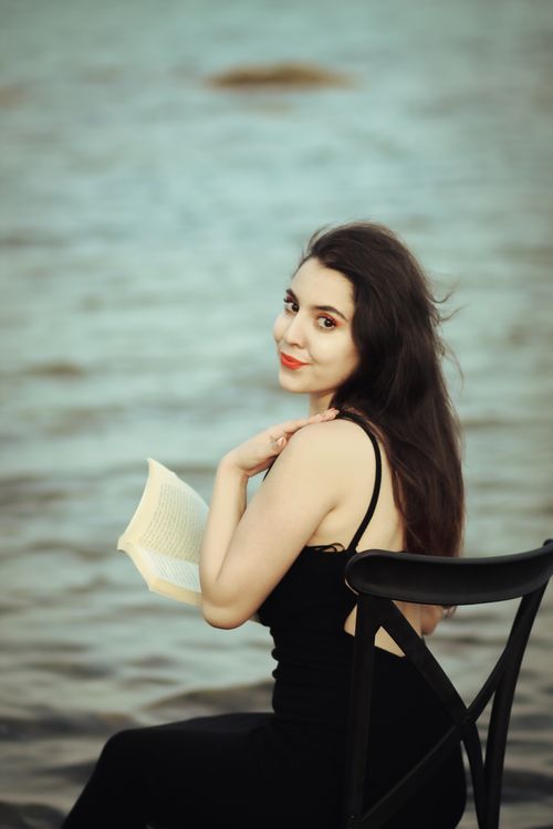 Free Woman sitting on a chair near water, holding a book and smiling. Stock Photo