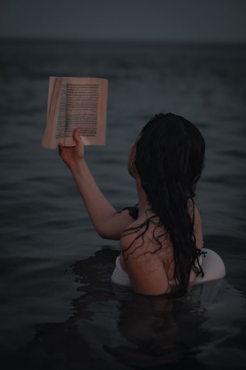 Free A young woman enjoys reading while partially submerged in water during twilight, creating a serene atmosphere. Stock Photo