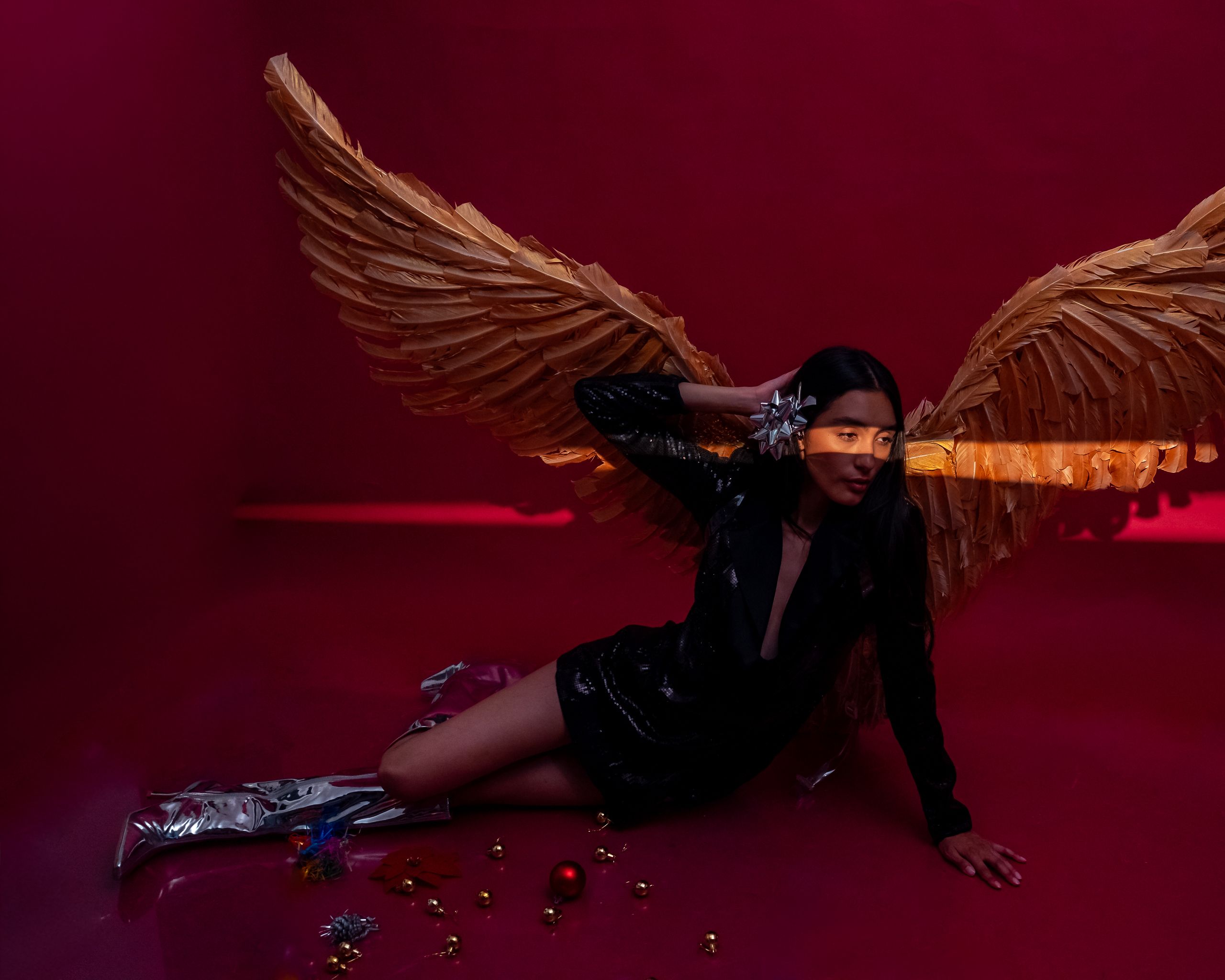 A woman poses elegantly with angel wings in a vibrant red background, exuding a festive, artistic charm.