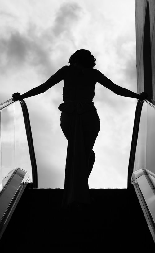 Free A dramatic silhouette of a woman ascending an escalator against a cloudy sky background. Stock Photo