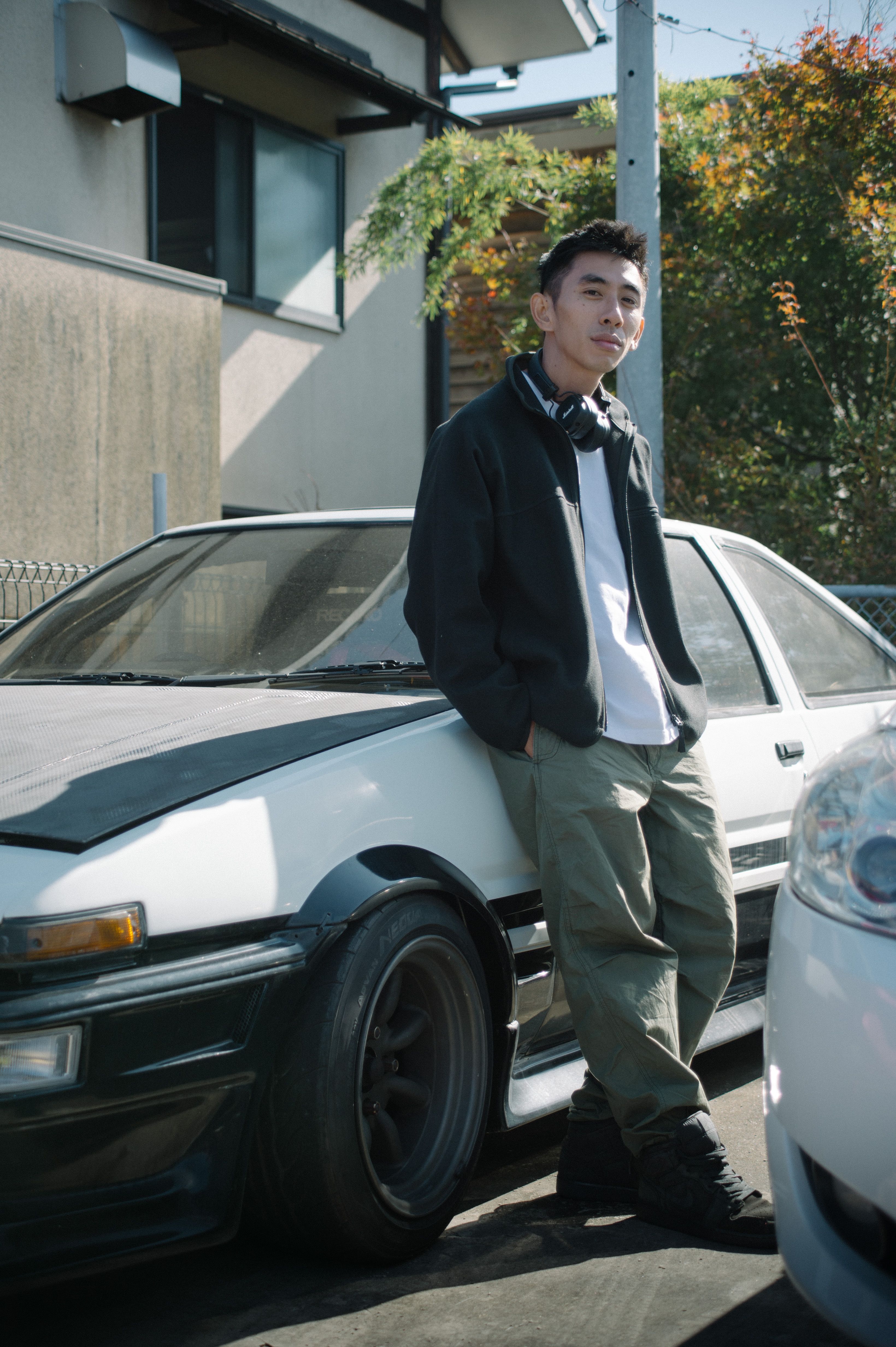 Young man in casual attire leans on a classic car in a Japanese neighborhood, showcasing style and urban culture.