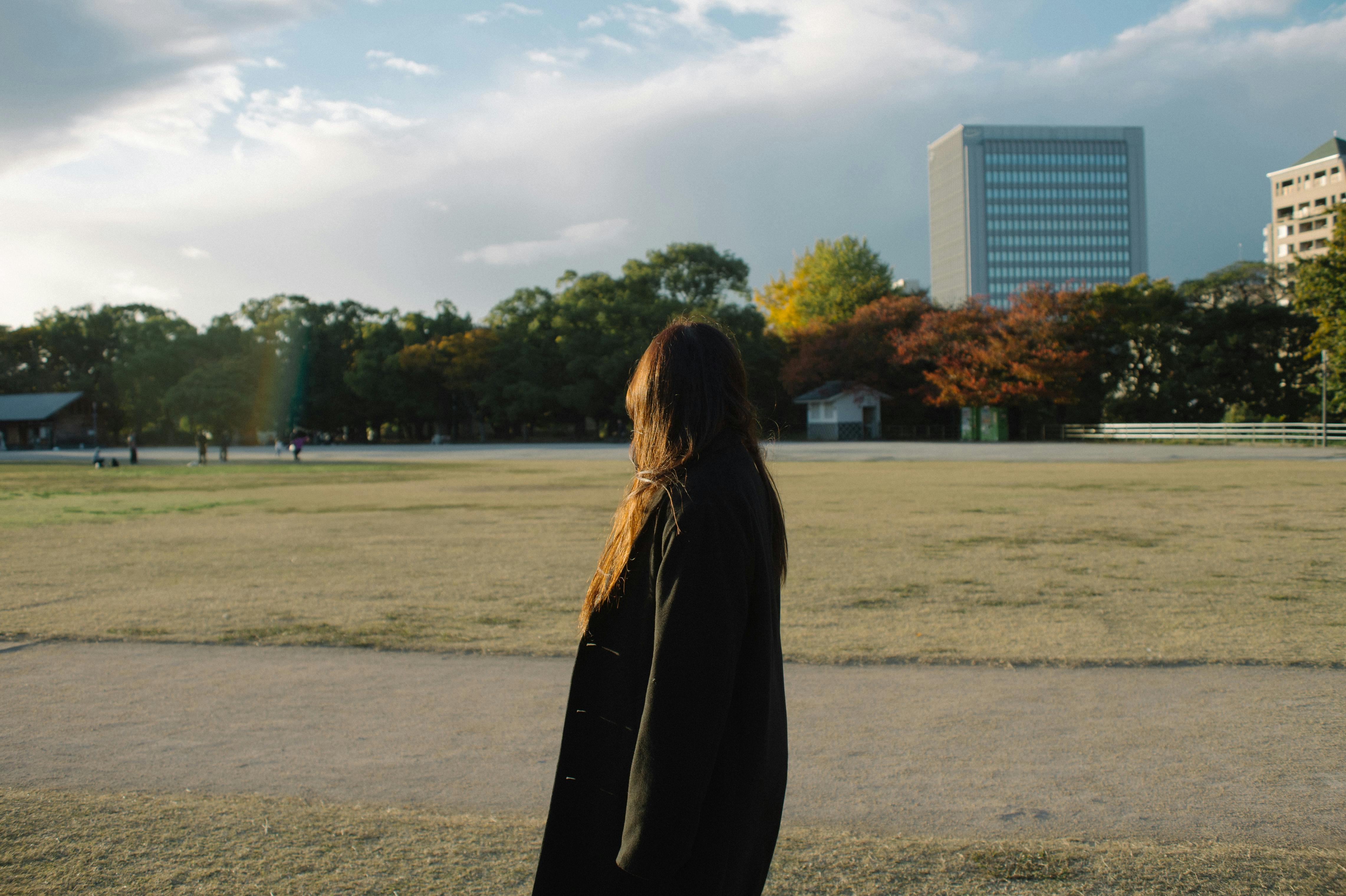 A person standing outdoors in a Japanese park with colorful autumn trees and a city skyline backdrop.