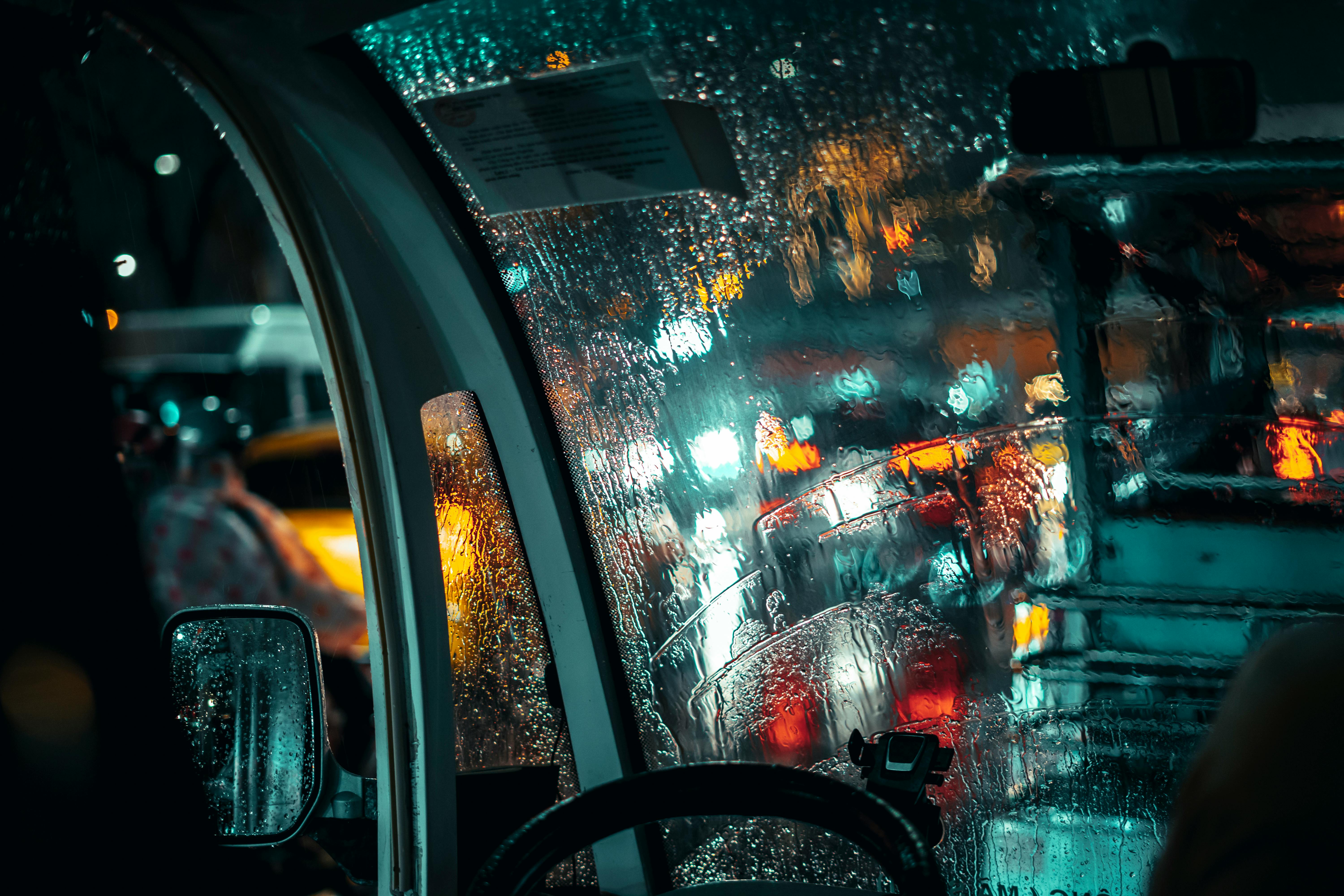 A moody view of city traffic through a rain-soaked vehicle window at night.