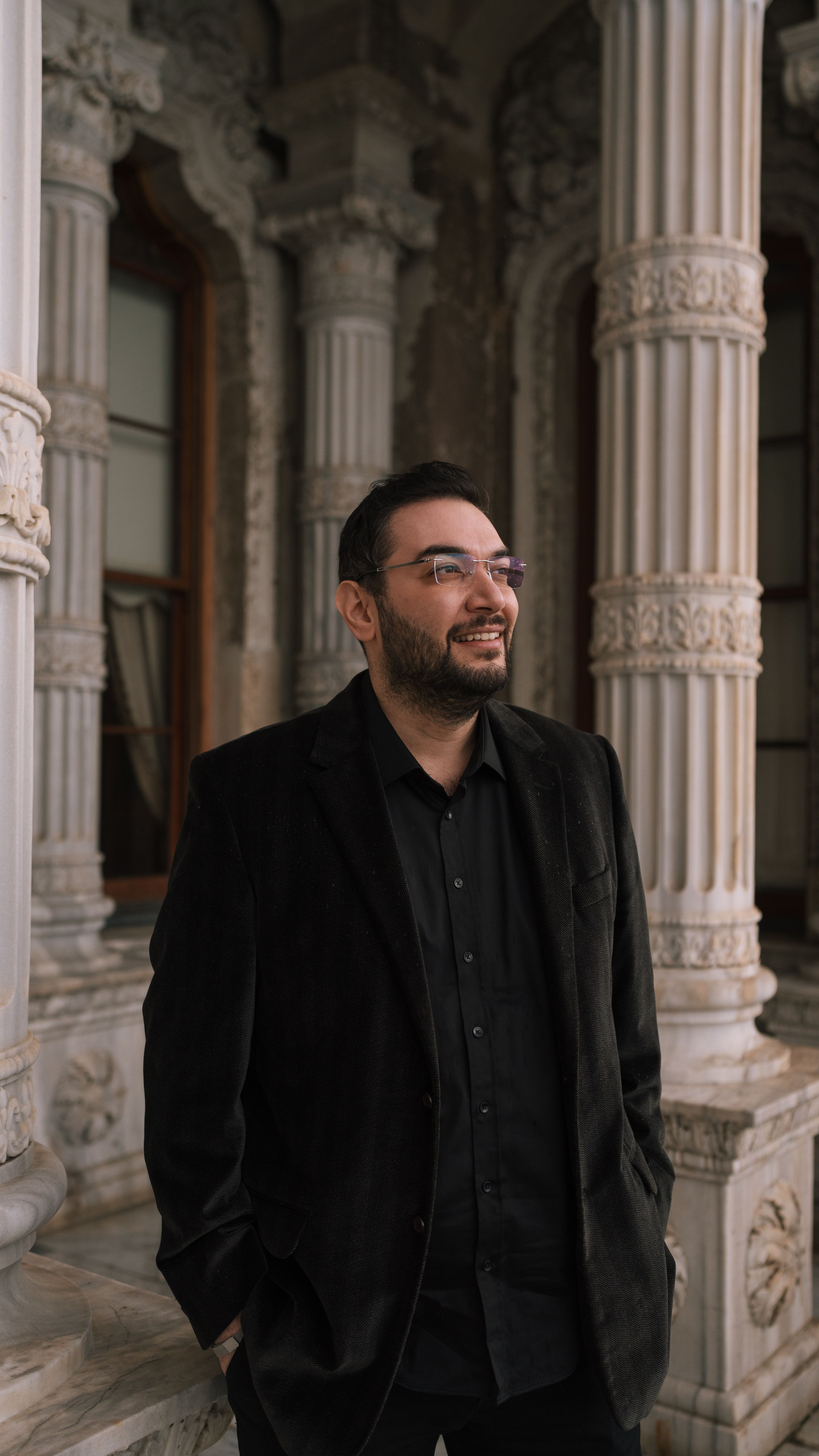 Man in dark suit standing by intricate columns, smiling.