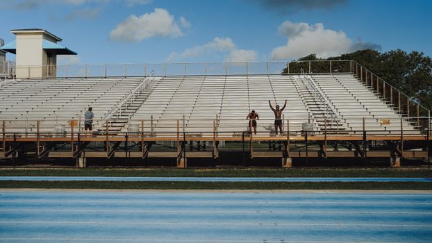 Two athletes and a coach in an empty stadium, preparing for a workout under a blue sky.