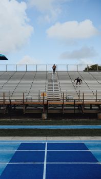 Two athletes perform training exercises on outdoor stadium bleachers under a clear sky.