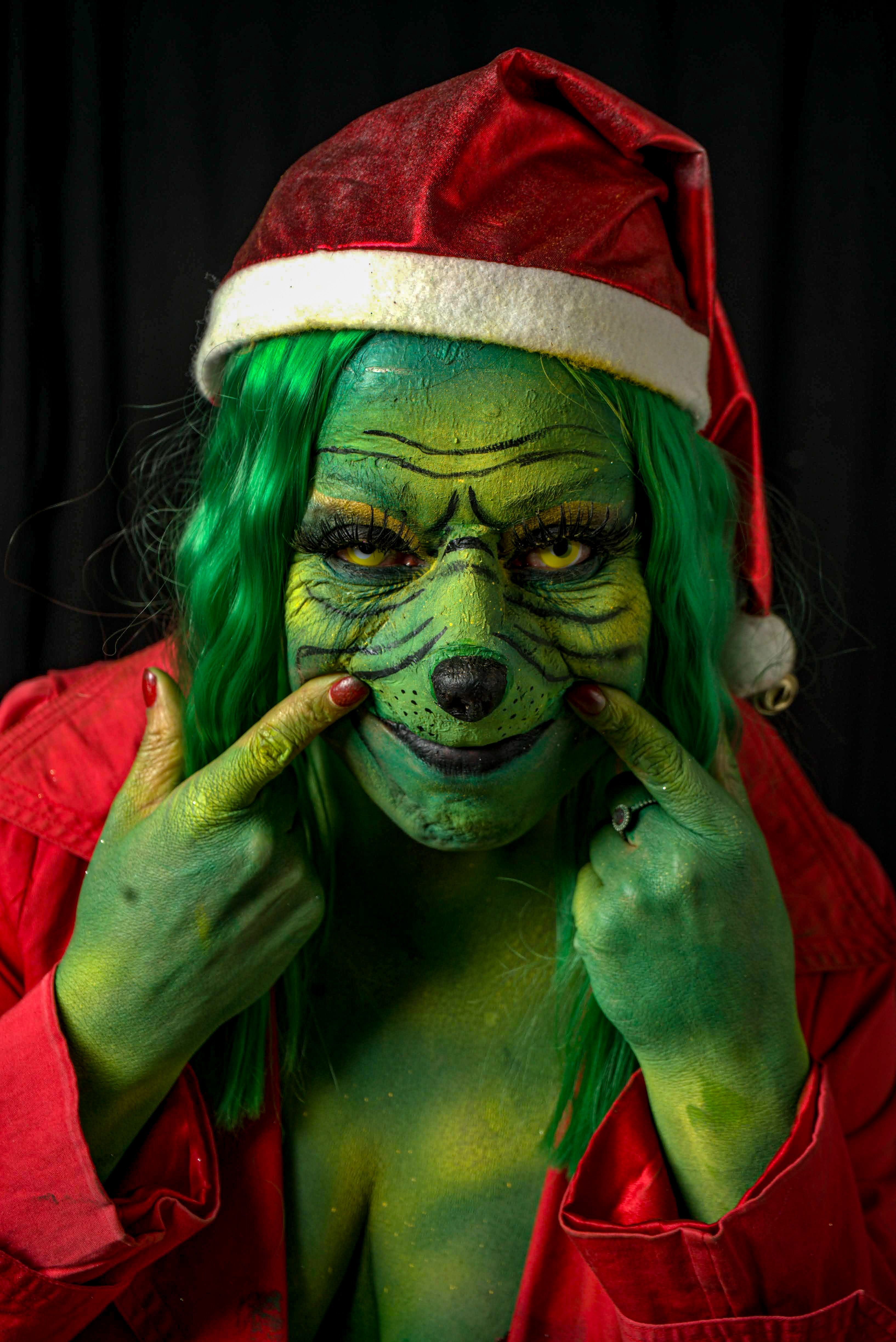Person dressed as green character with Santa hat and red coat, indoors.