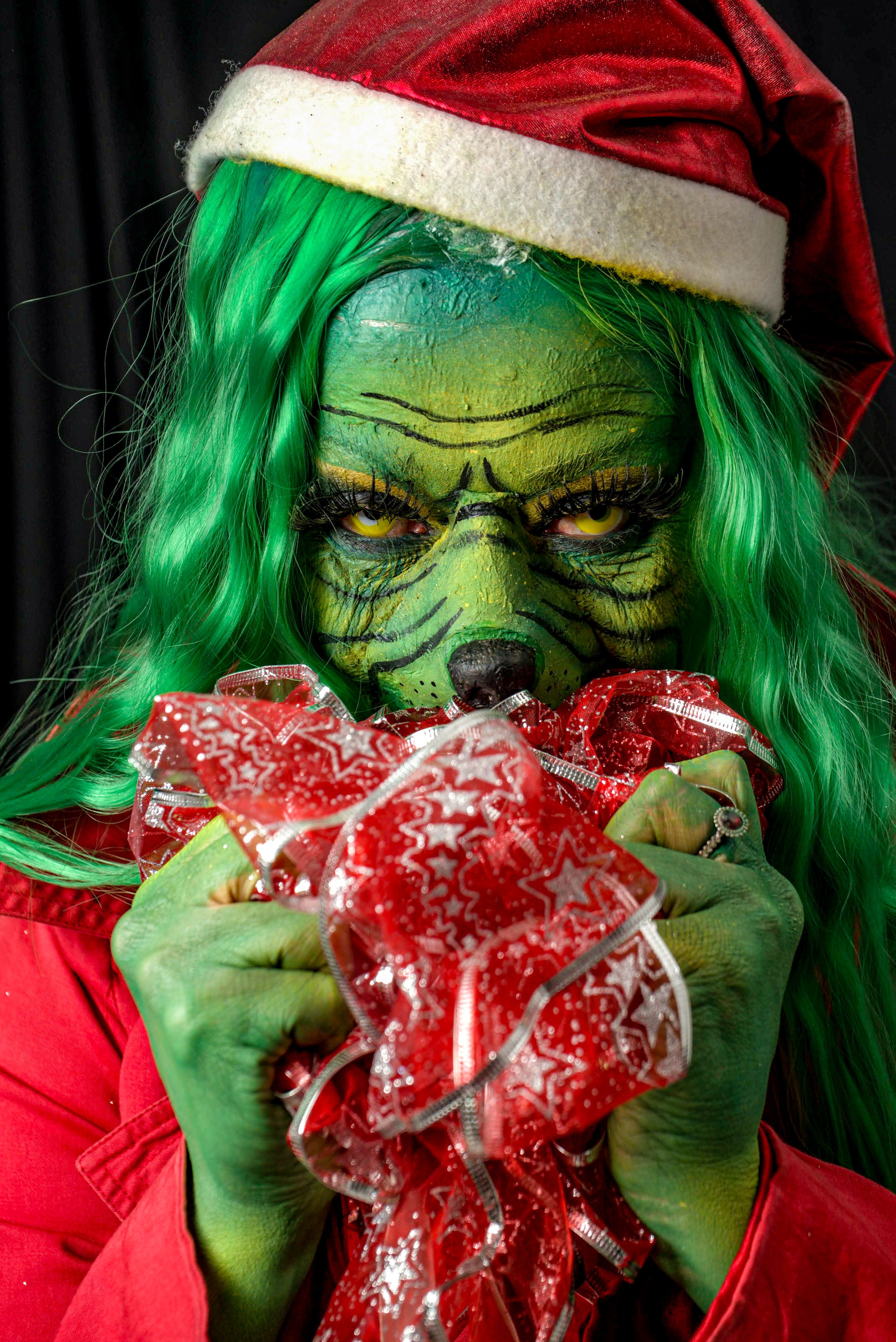 A person dressed as the Grinch, wearing a Santa hat, clutching a red holiday ribbon.
