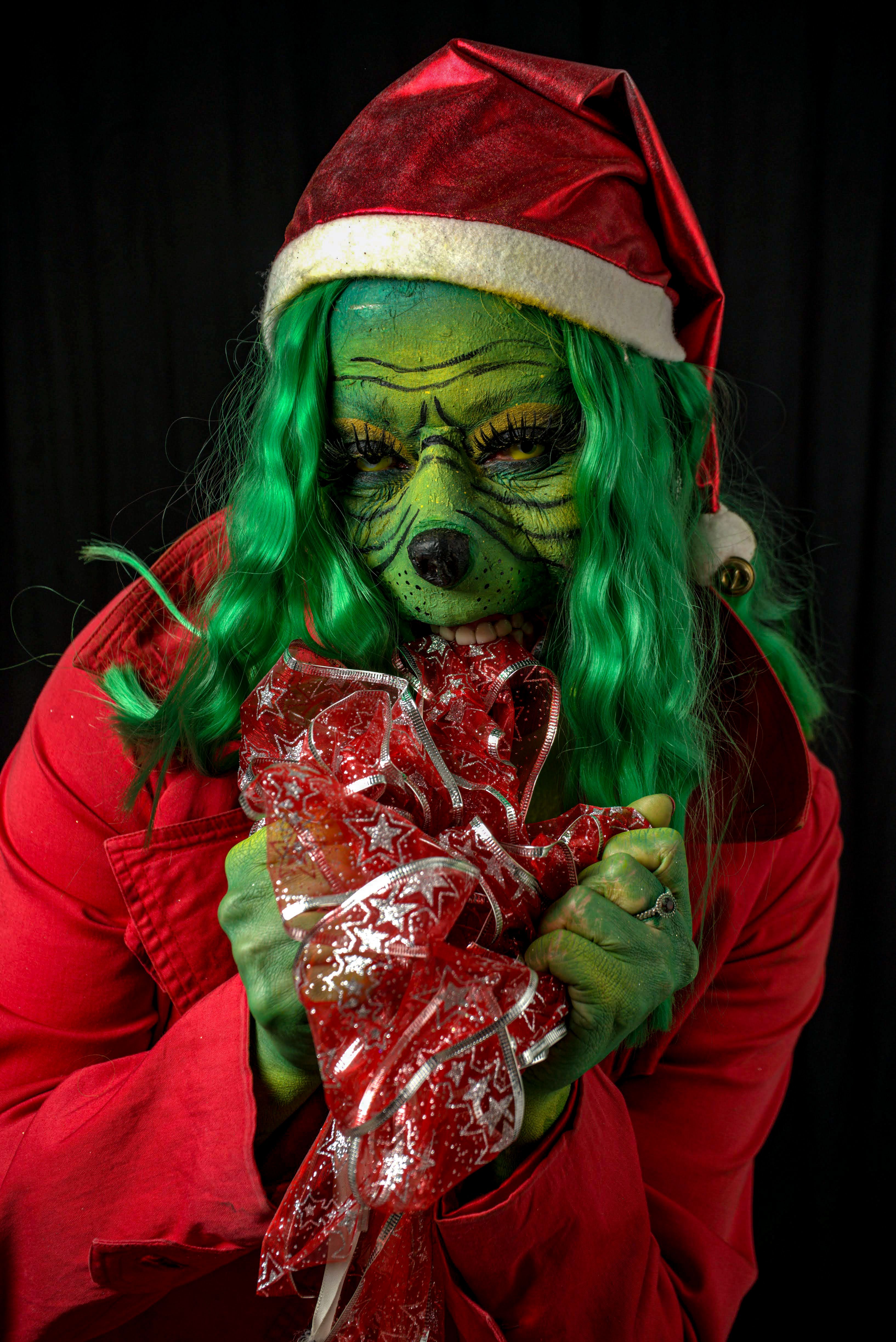 Person in Grinch costume holding a festive Christmas gift with green face paint and red attire.