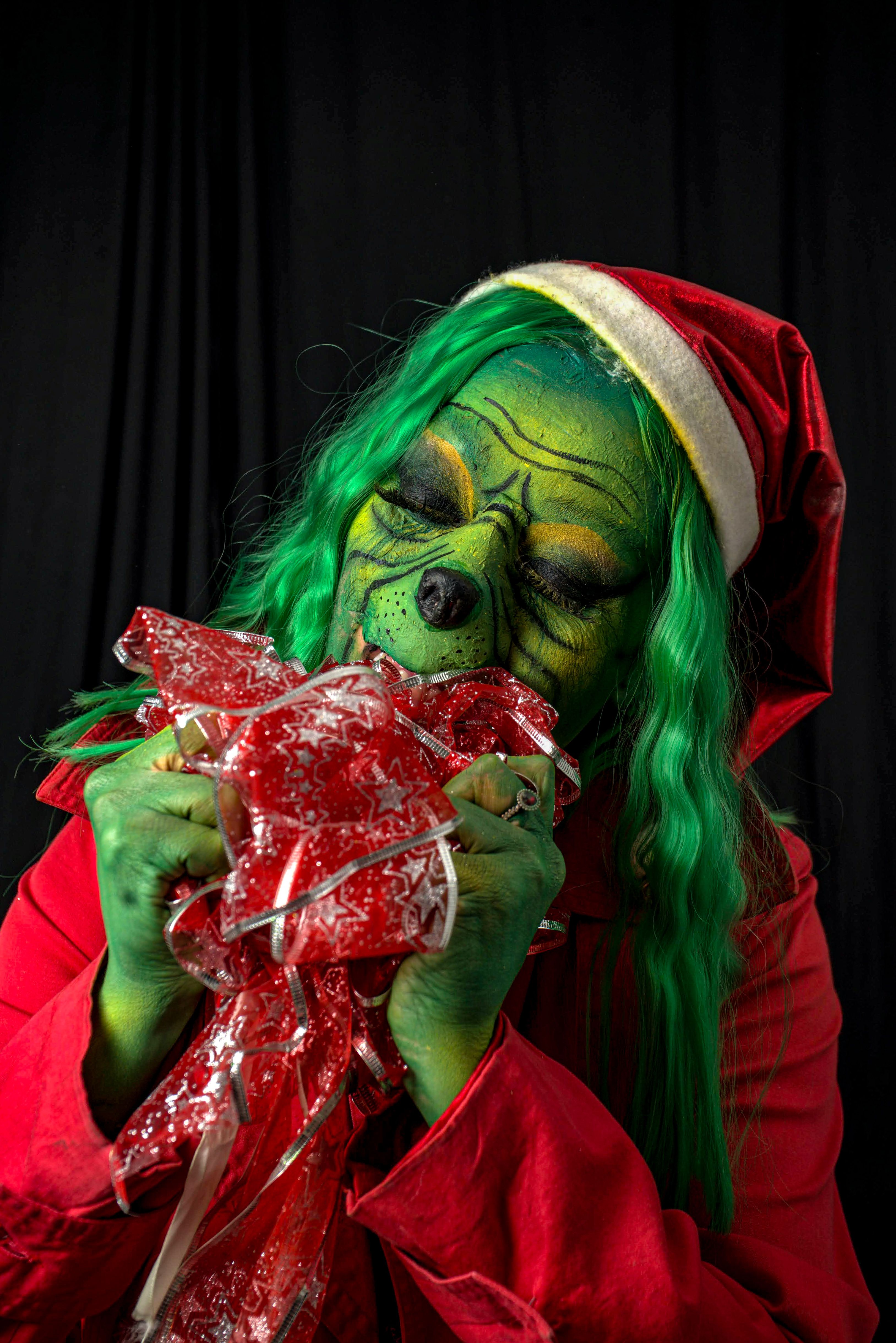 A person dressed as the Grinch, clutching a festive wrapped gift with a joyful expression.
